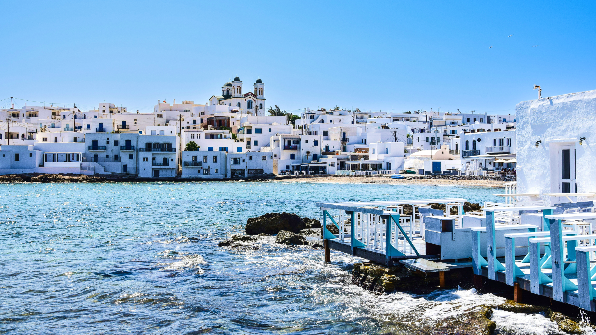 Naoussa village in Paros with pristine whitewashed buildings and church dominating the skyline - a popular destination during Greek Cyclades island hopping with traditional blue accents and waterfront dining visible from the harbor