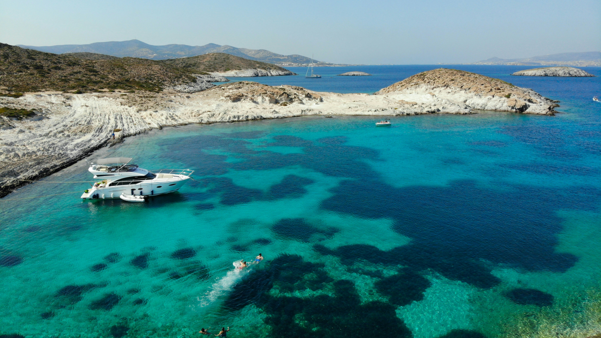 Luxury motor yacht anchored in crystal-clear turquoise waters near Antiparos during Greek Cyclades island hopping - aerial view showing swimmers enjoying secluded cove with distinctive white marble rock formations and shallow reefs visible beneath transparent water