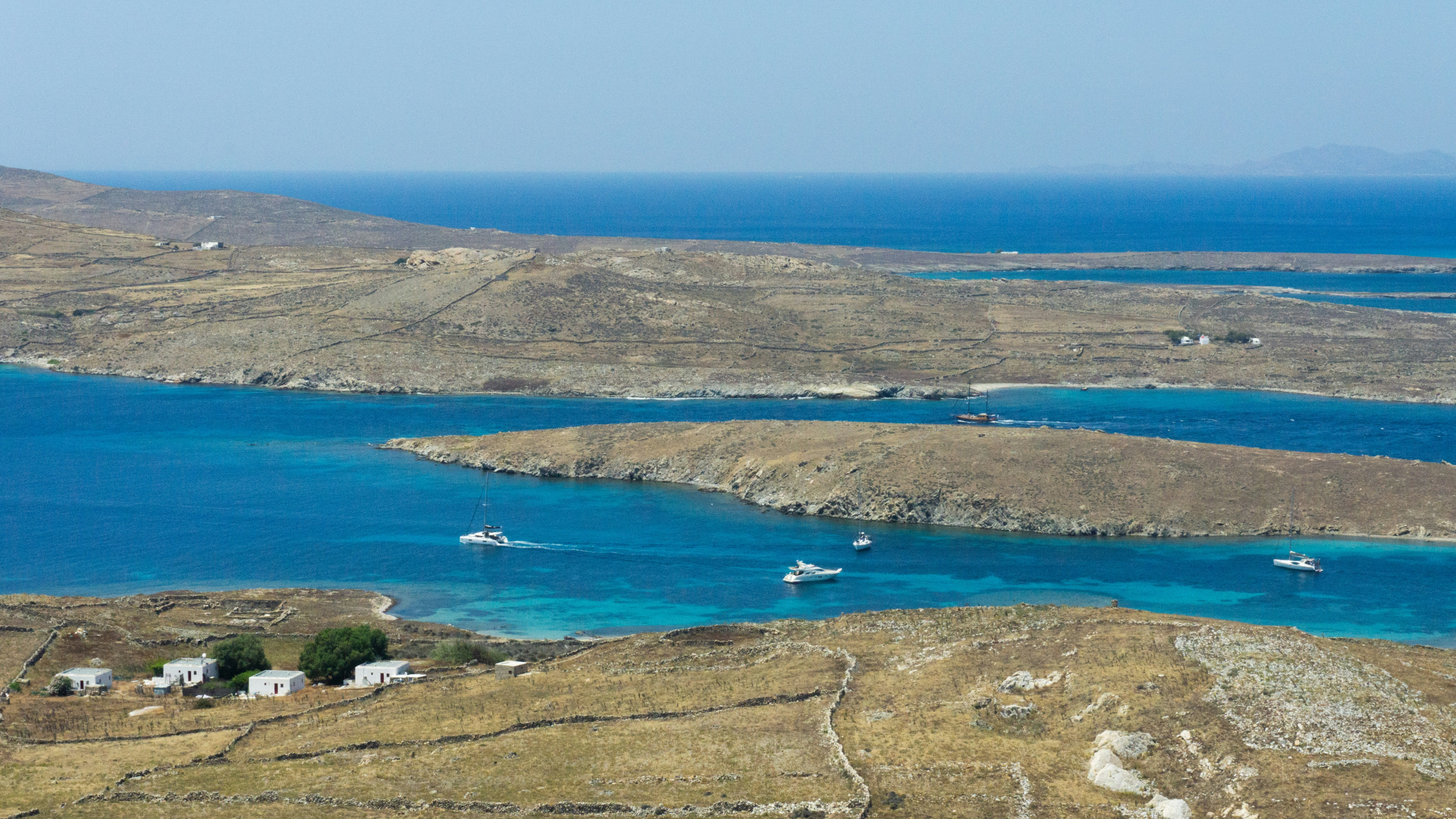 Luxury yachts sailing through brilliant turquoise waters during Greek Cyclades island hopping near Delos - aerial view showing the rugged island landscape with characteristic white houses and stunning blue bays perfect for anchoring