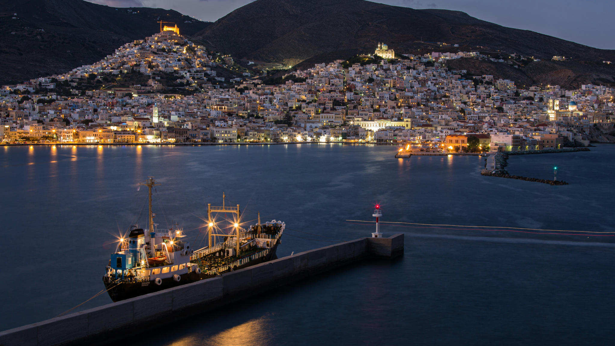 Evening view of Ermoupoli in Syros illuminated at twilight - a spectacular final stop for Greek Cyclades island hopping with its amphitheater-like city climbing hillsides, neoclassical architecture, and harbor with lighthouse visible as yachts return from sailing