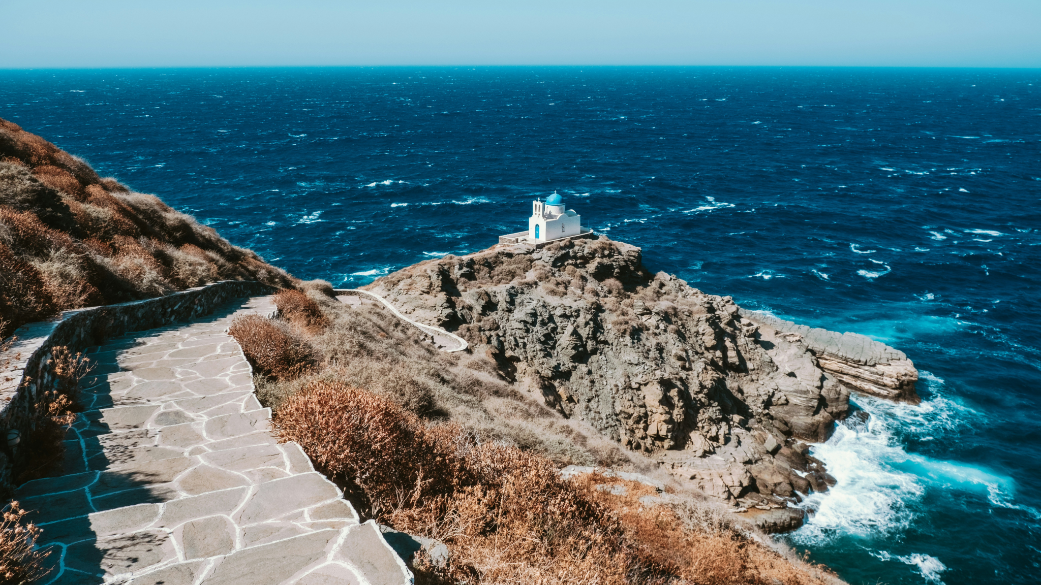 Iconic blue-domed chapel perched on rocky promontory in Sifnos overlooking deep azure Aegean waters - a spectacular sight during Greek Cyclades island hopping with traditional stone pathway leading to this solitary church against the vast sea