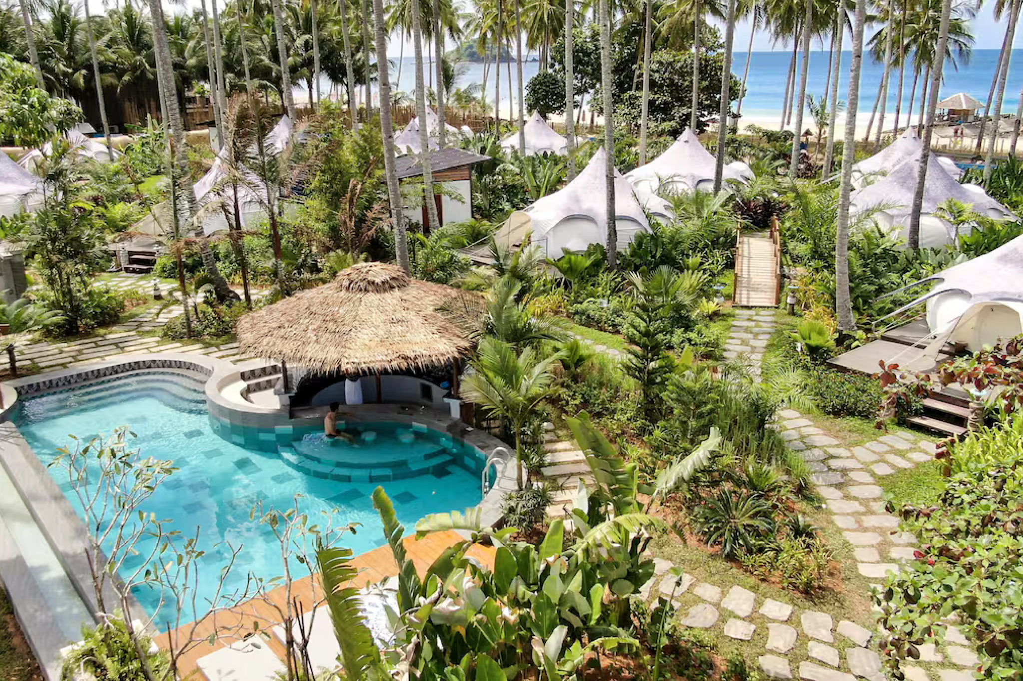 Aerial view of Nacpan Beach Glamping resort in El Nido Palawan, with luxury tents surrounded by palm trees, a pool with a thatched swim-up bar, and white-sand beach access — one of the best beach resorts in El Nido Palawan.