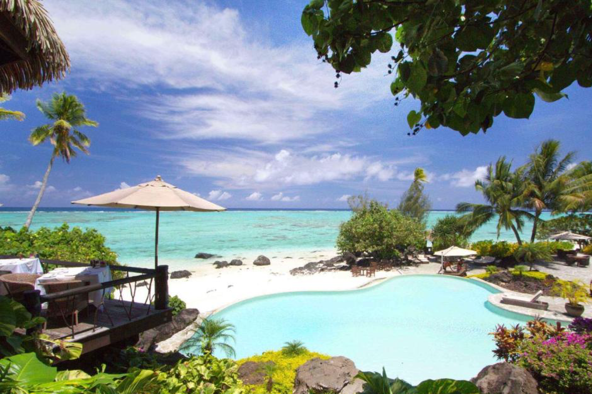 Panoramic view of Pacific Resort Aitutaki’s infinity pool and lagoonfront terrace surrounded by tropical gardens and white sand, one of the best Aitutaki resorts for luxury travelers.