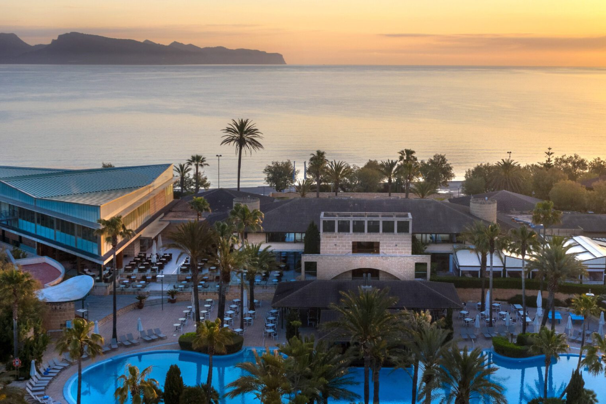 Aerial view of Hipotels Mediterráneo Hotel in Sa Coma at sunrise, featuring a serene beachfront, palm trees, and a large pool — one of the best beach resorts in Mallorca for adults-only stays.