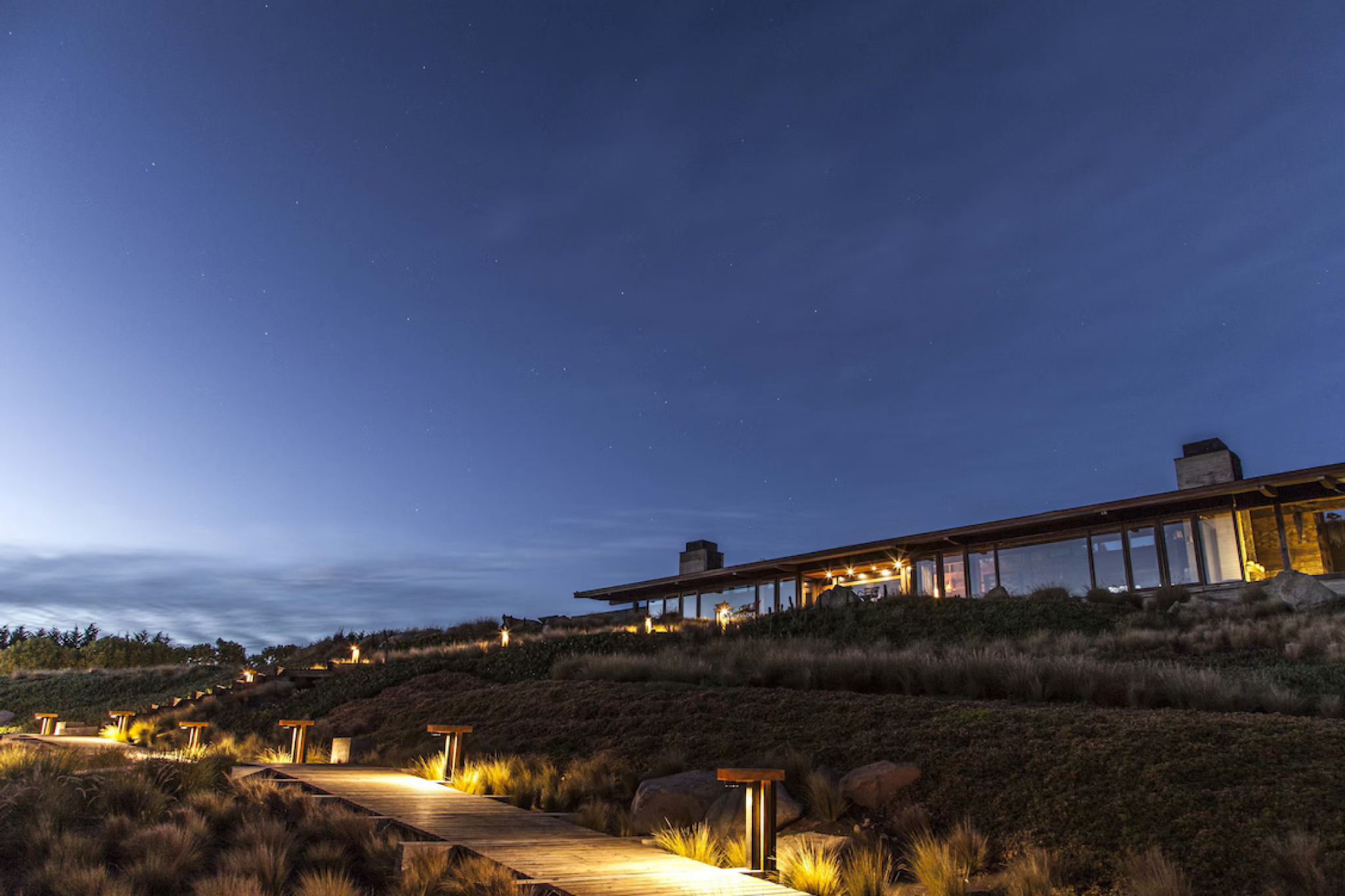A dramatic twilight view of Hotel Alaia in Punta de Lobos, with its minimalist wooden structures glowing softly against the night sky and a winding illuminated boardwalk leading toward the surfside cliffs — a rugged coastal setting that reflects why the property ranks among the best beach resorts in South America in 2025.