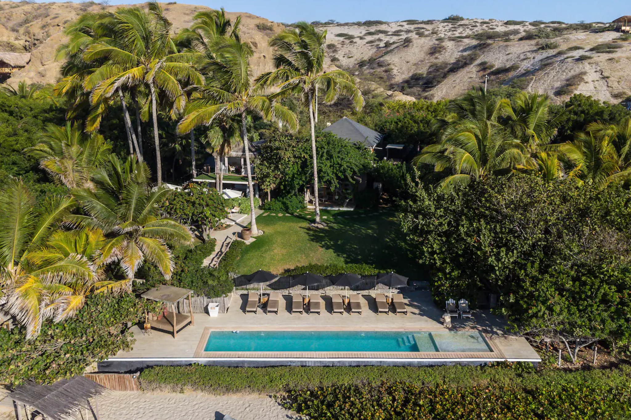 An aerial view of KiChic in Mancora showing its beachfront pool lined with shaded loungers, surrounded by lush palm trees and backed by golden desert cliffs — a serene adults-only sanctuary highlighted in the best beach resorts in South America in 2025 for its peaceful, bohemian atmosphere.