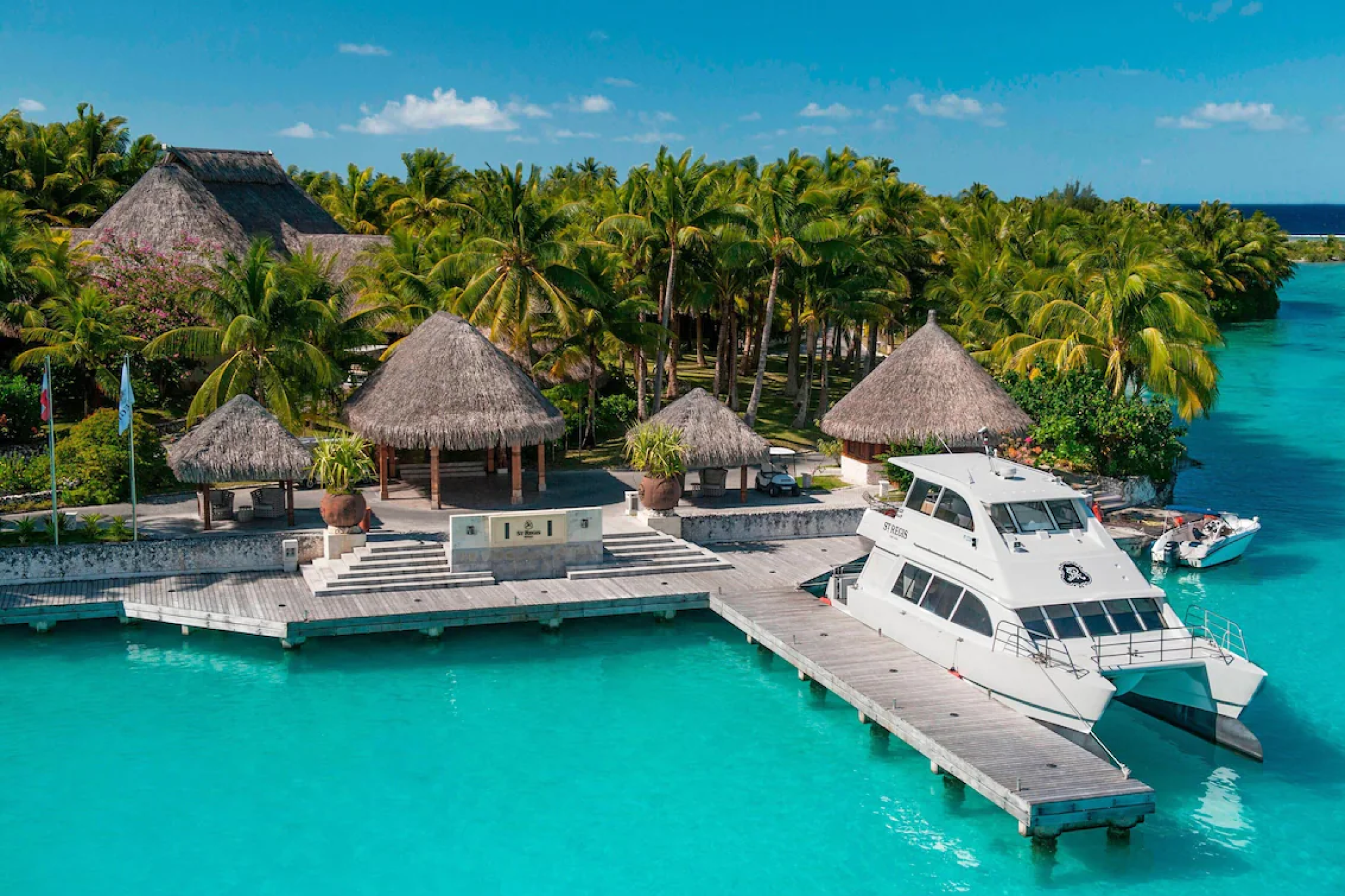 The dock and thatched-roof pavilions of St. Regis Bora Bora Resort beside a bright turquoise lagoon, with palm gardens and anchored yachts — a signature setting of one of the best beach resorts in the South Pacific in 2025.