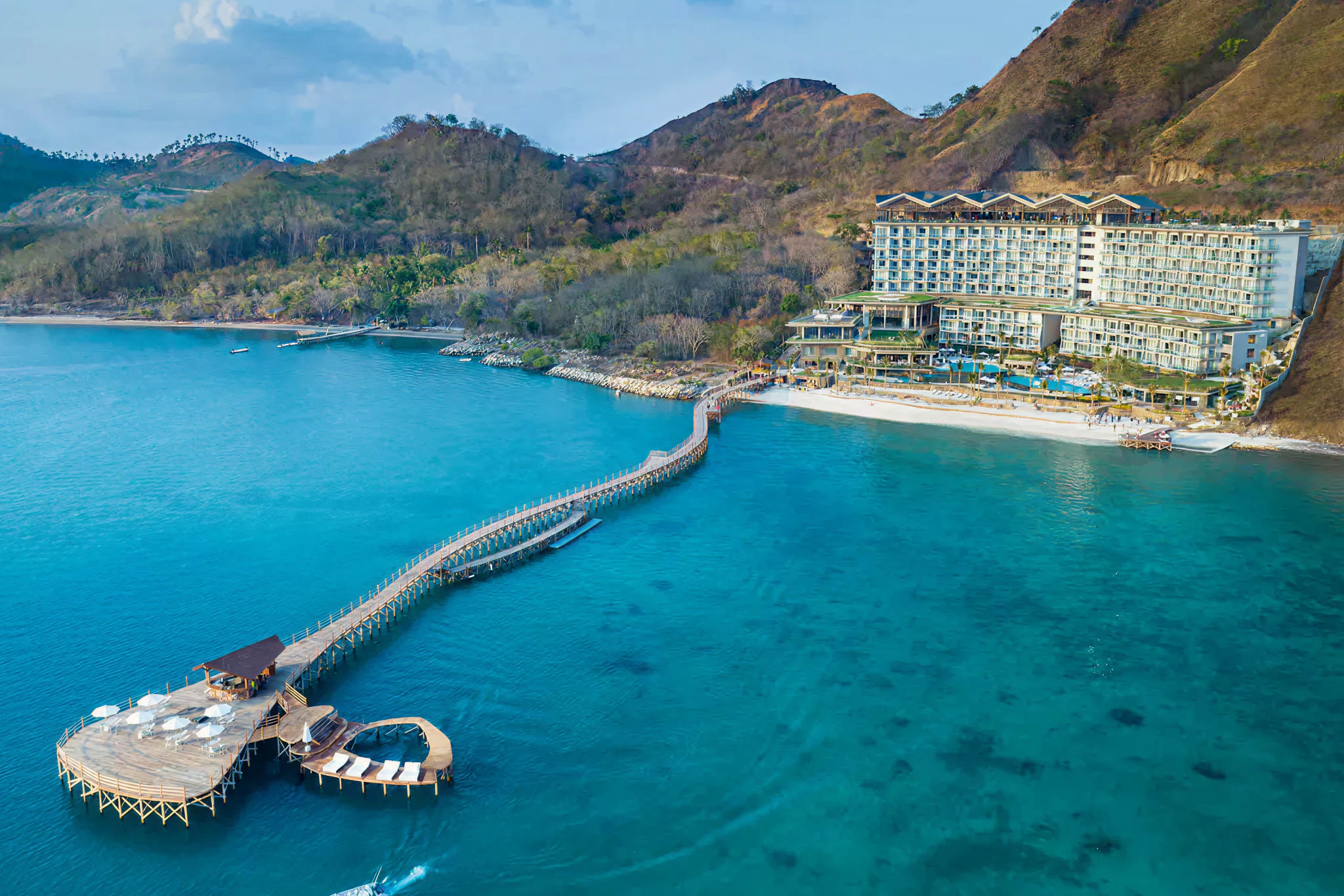 An expansive coastal shot of AYANA Komodo Waecicu Beach, highlighted as one of the best beach resorts in Southeast Asia in 2025, showcasing its dramatic beachfront location, long wooden jetty, and crystal-clear blue waters facing Komodo National Park.