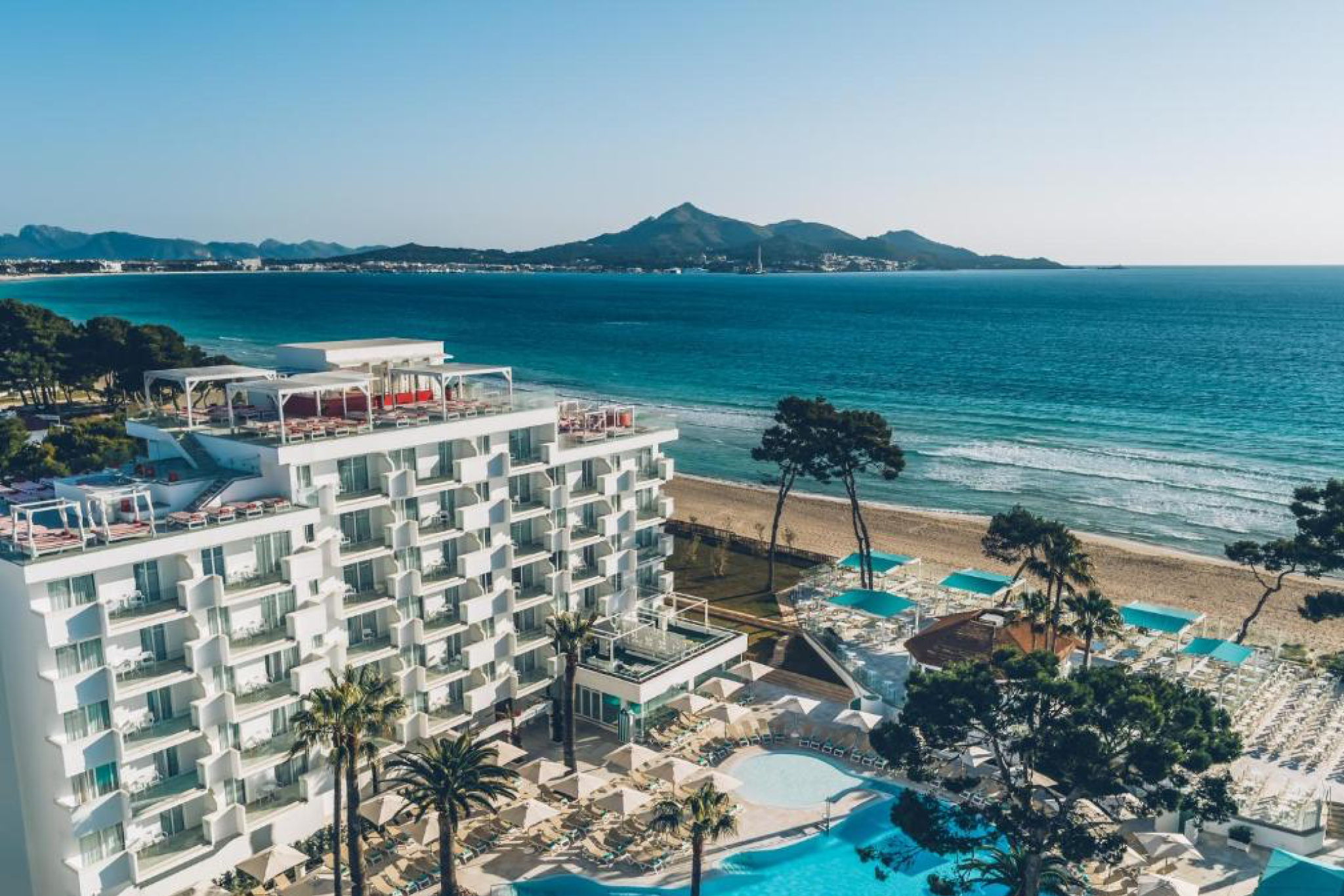 Overhead shot of Iberostar Waves Alcudia Park showcasing its modern white façade, expansive terraces, and beachfront pools directly beside Alcudia’s clear blue waters — part of the best beach resorts in Spain for families and couples.