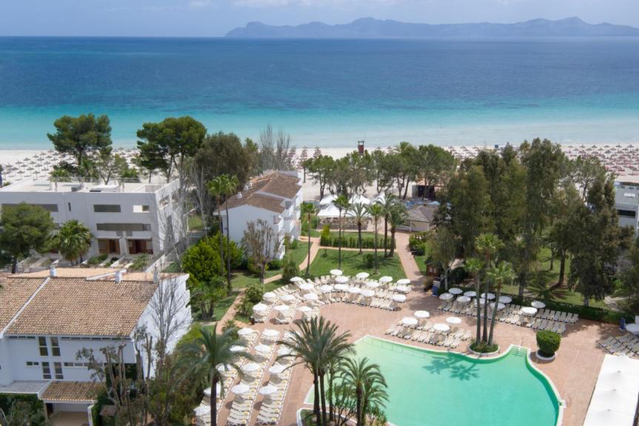 Bright aerial image showing Iberostar Waves Ciudad Blanca with whitewashed buildings, palm-lined gardens, and a large pool facing Alcudia Bay’s soft golden sand — a coastal haven recognized as one of the best beach resorts in Spain.