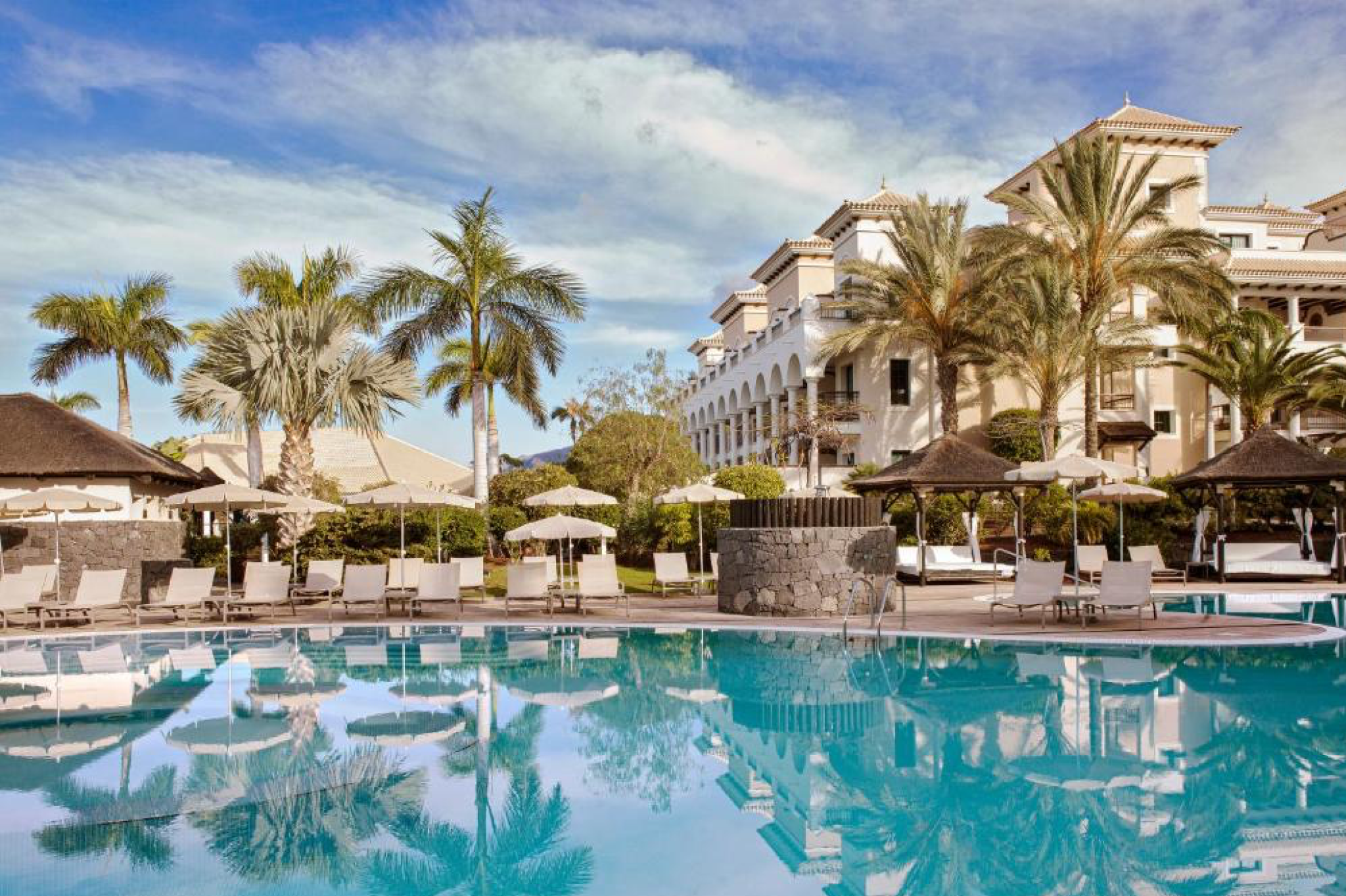 Elegant resort scene of RedLevel at Gran Meliá Palacio de Isora, featuring palm trees reflected in a crystal-blue infinity pool beneath a clear Tenerife sky — an exclusive destination among the best beach resorts in Spain.