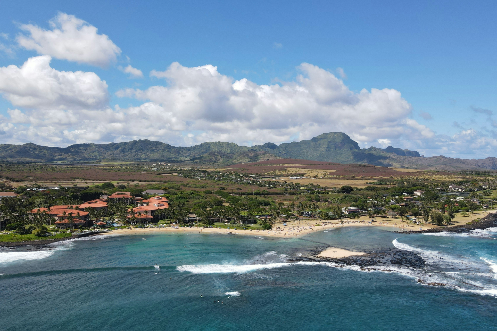 An aerial view of Poipu Beach on Kauai’s south shore, showcasing calm turquoise waters, golden sand, and the Grand Hyatt—one of the best beaches in Kauai for families and year-round sun.