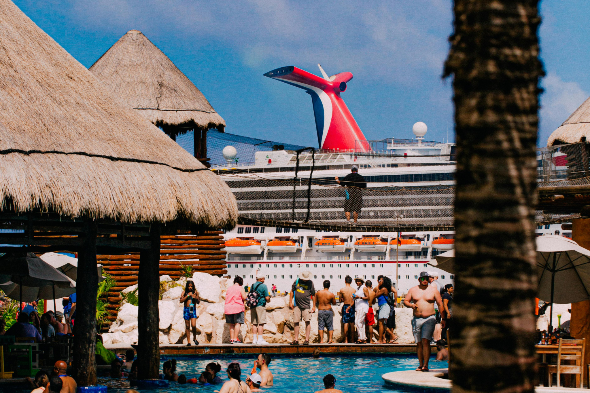 Vibrant view of cruise passengers enjoying a tropical beach with thatched-roof shelters, lifeboats, and a docked Carnival cruise ship, illustrating Cozumel as a top Caribbean port and one of the best cruise ports in 2025.