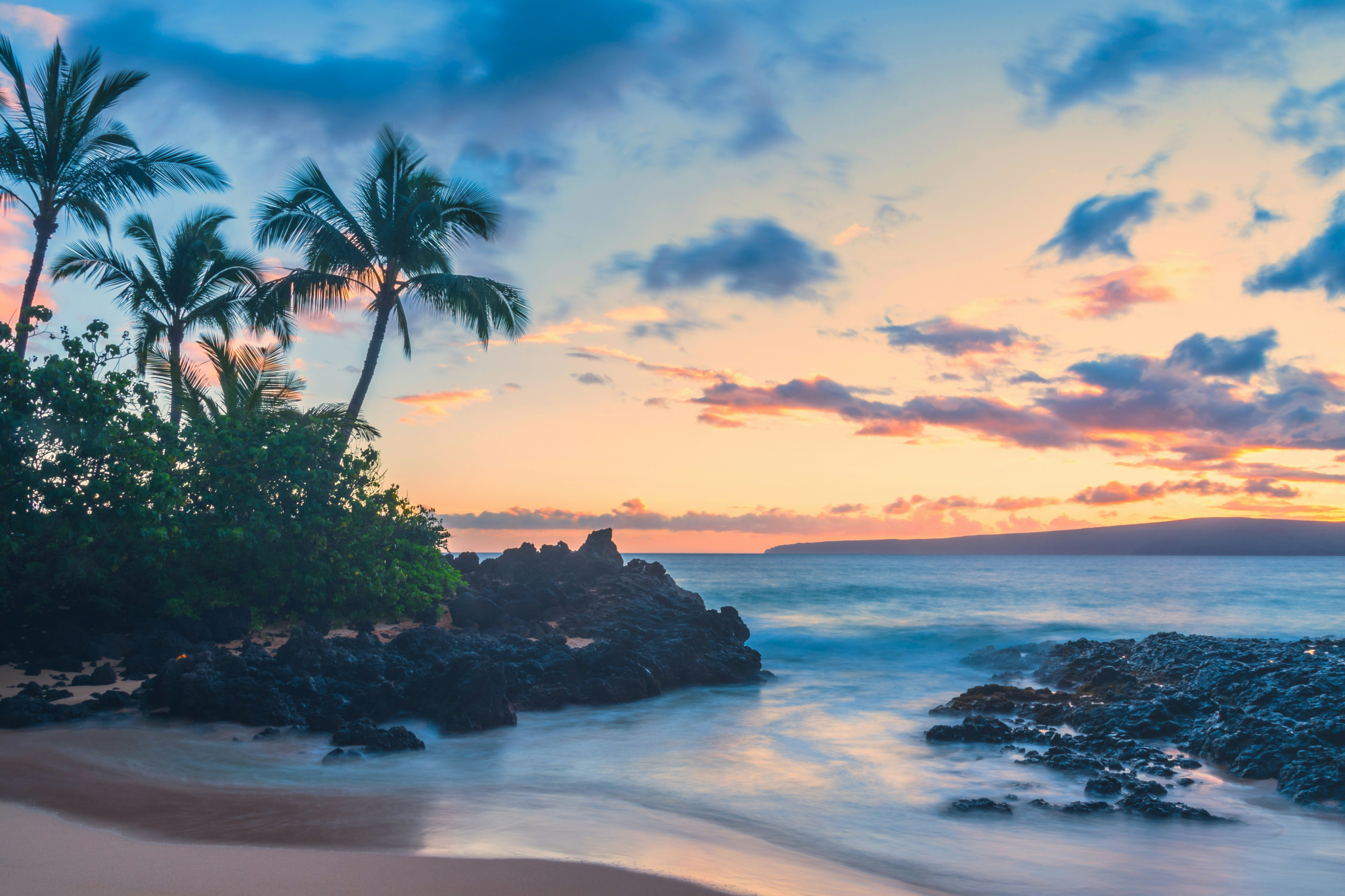 Sunset shoreline in Maui with palm silhouettes, lava rock formations, and soft pastel skies reflecting over a gently moving Pacific Ocean — highlighting the natural beauty that makes Maui one of the best islands in the world in 2025.