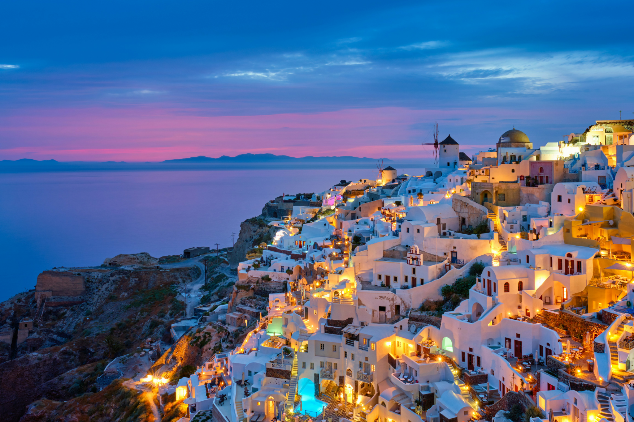Twilight view of Santorini’s iconic whitewashed cliffside homes glowing with warm lights above a calm Aegean Sea, set beneath a purple-pink sunset — an unmistakable scene from one of the best islands in the world in 2025.