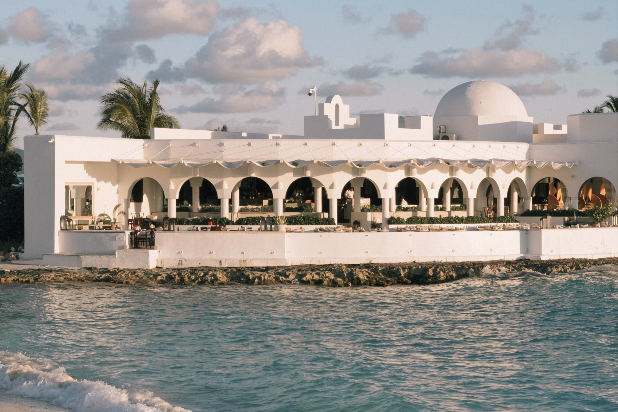 Seafront dining terrace at Cap Juluca, A Belmond Hotel, perched above Maundays Bay at golden hour with Mediterranean arches and ocean waves below.