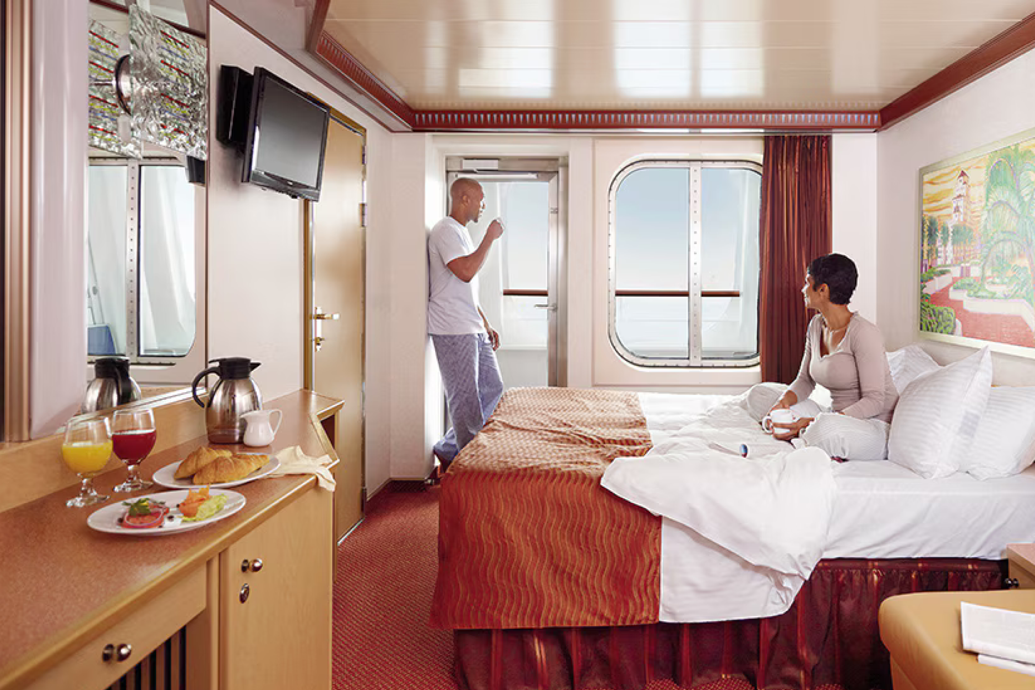 Interior view of a Carnival Firenze balcony stateroom with a couple enjoying ocean views, featuring breakfast setup and warm-toned furnishings.