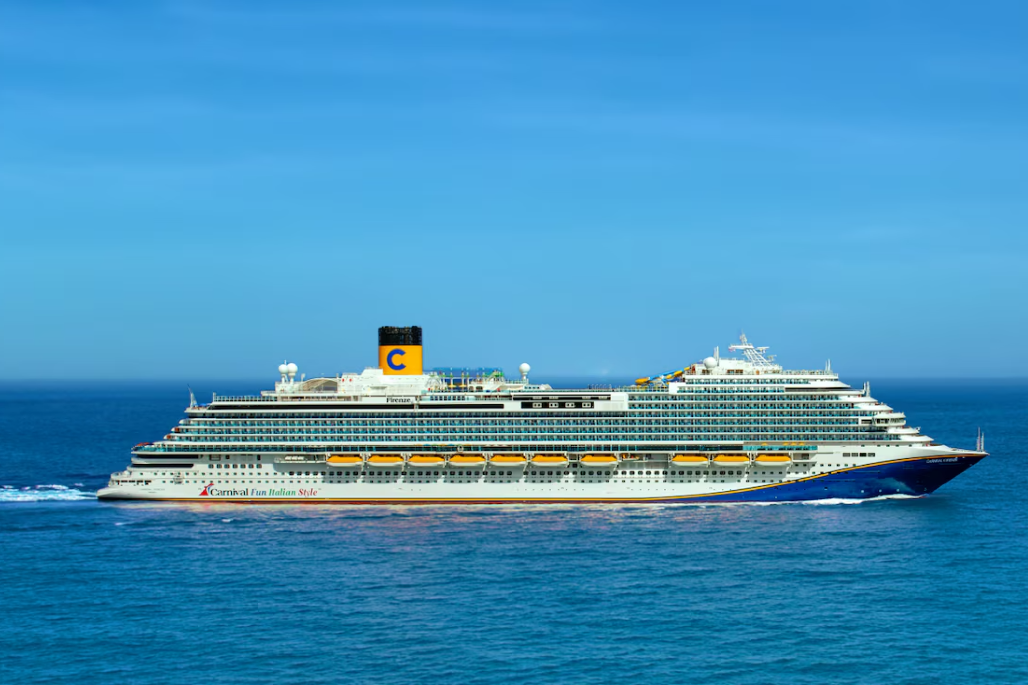 Carnival Firenze sailing at sea under clear blue skies, displaying the full length of the ship, lifeboats, and signature Carnival funnel.