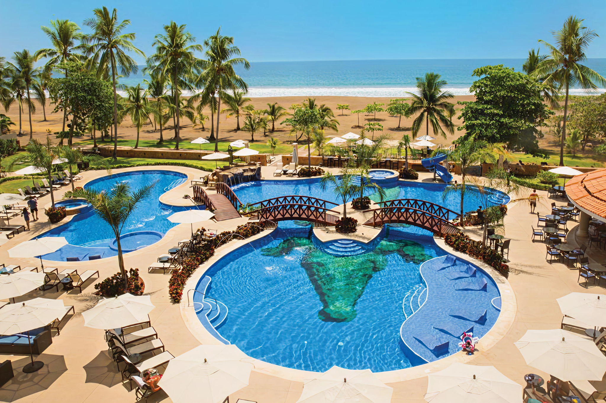 Aerial view of the main pool area at Crocs Resort & Casino with bridges, palm trees, loungers, and Jacó Beach in the background.