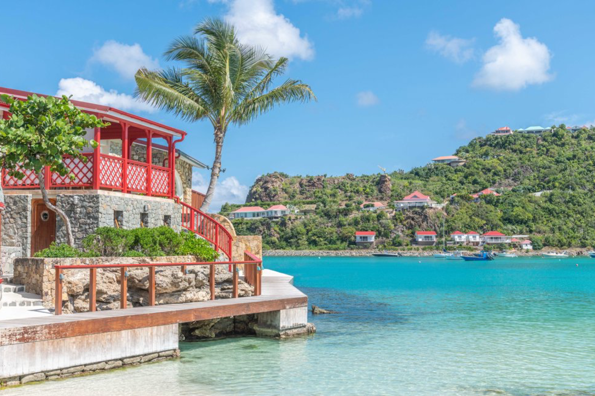 Waterfront stone building with red balcony at Eden Rock St. Barths, surrounded by clear blue sea and lush green hills.