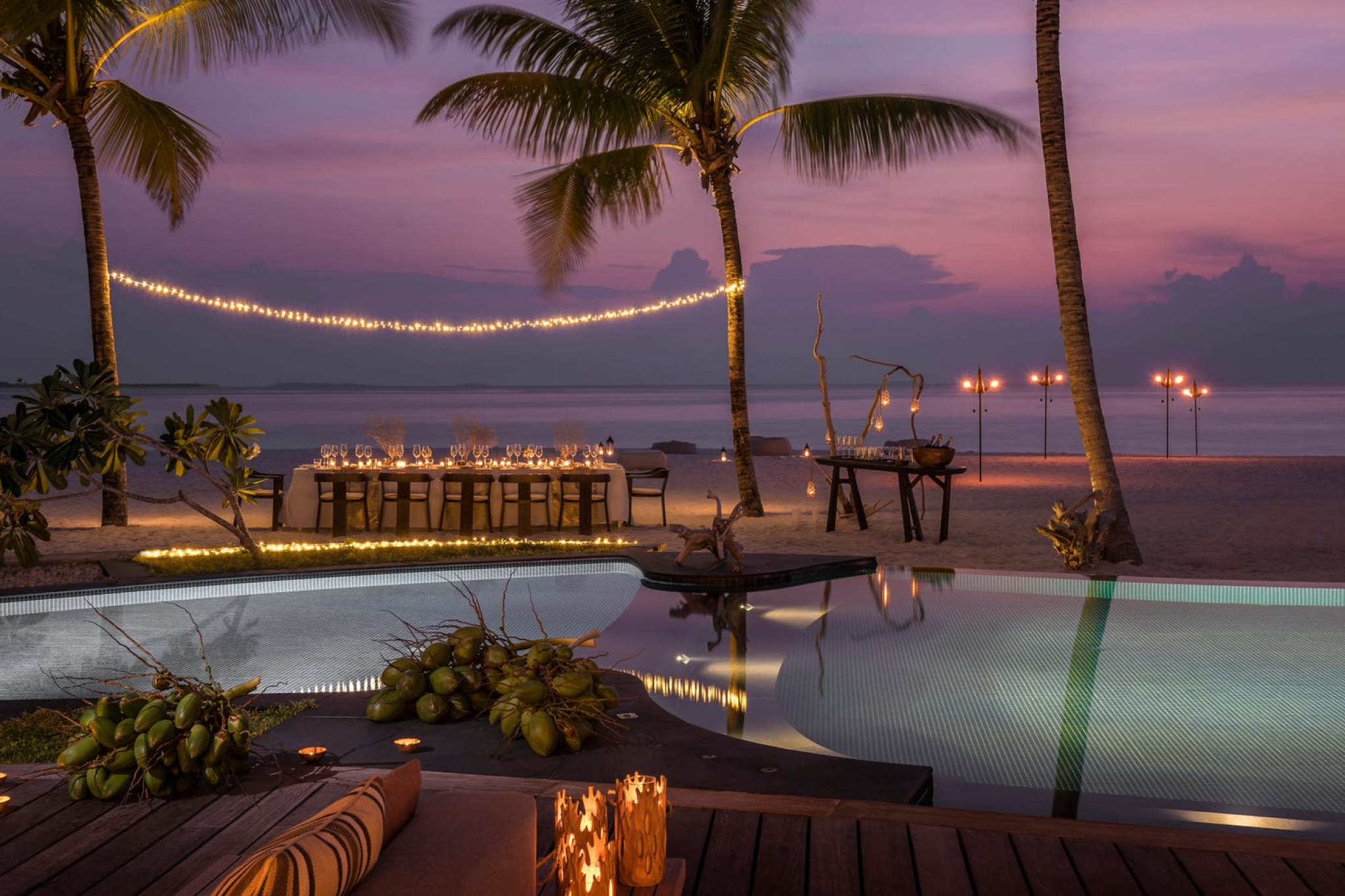 Private beachfront dinner setup under palm trees at Voavah, lit with string lights and candles at dusk.