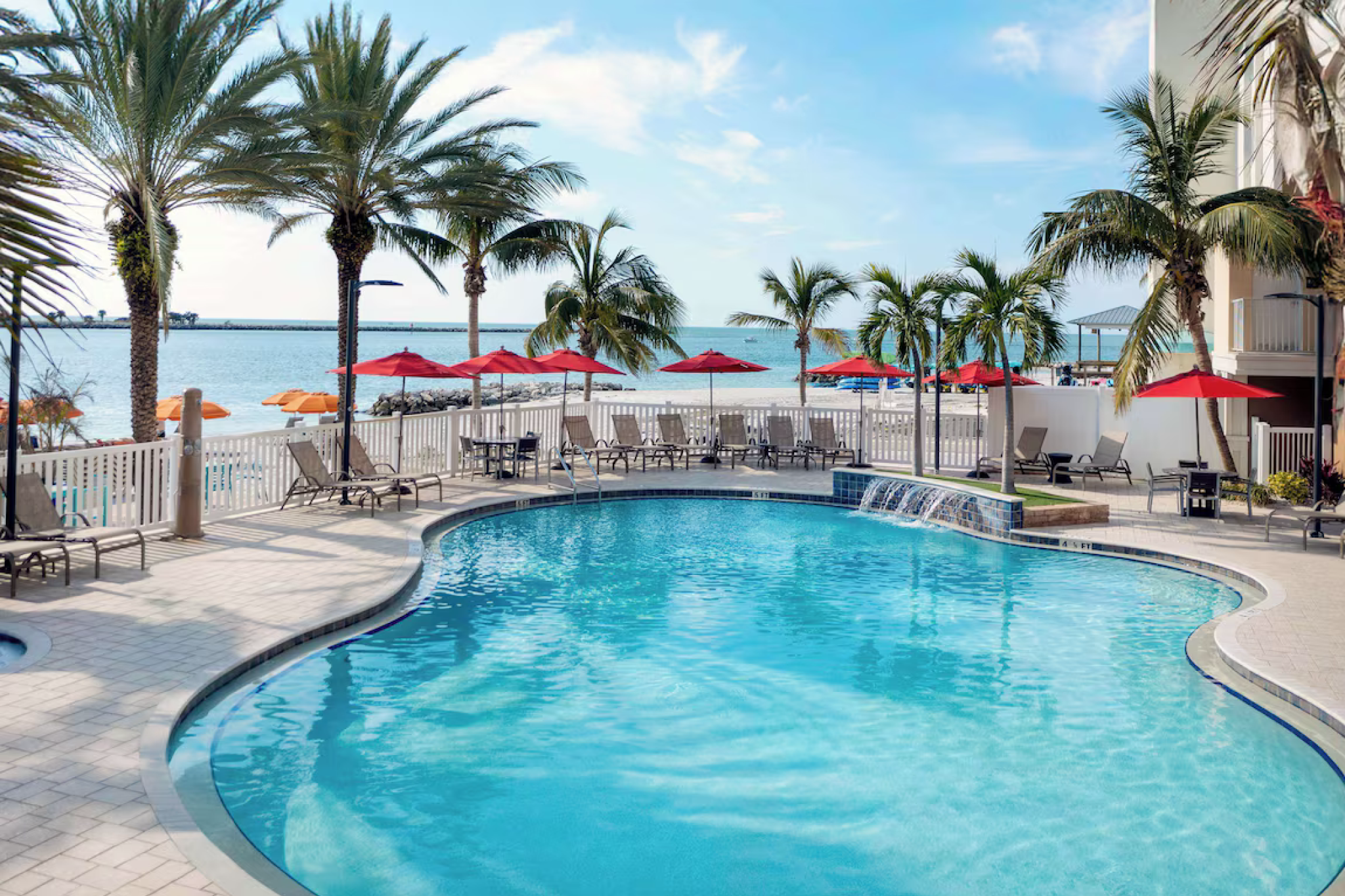 Beachfront pool at Hampton Inn & Suites Clearwater Beach featuring palm trees, red umbrellas, and lounge chairs with views of the white sand shoreline and Gulf waters.