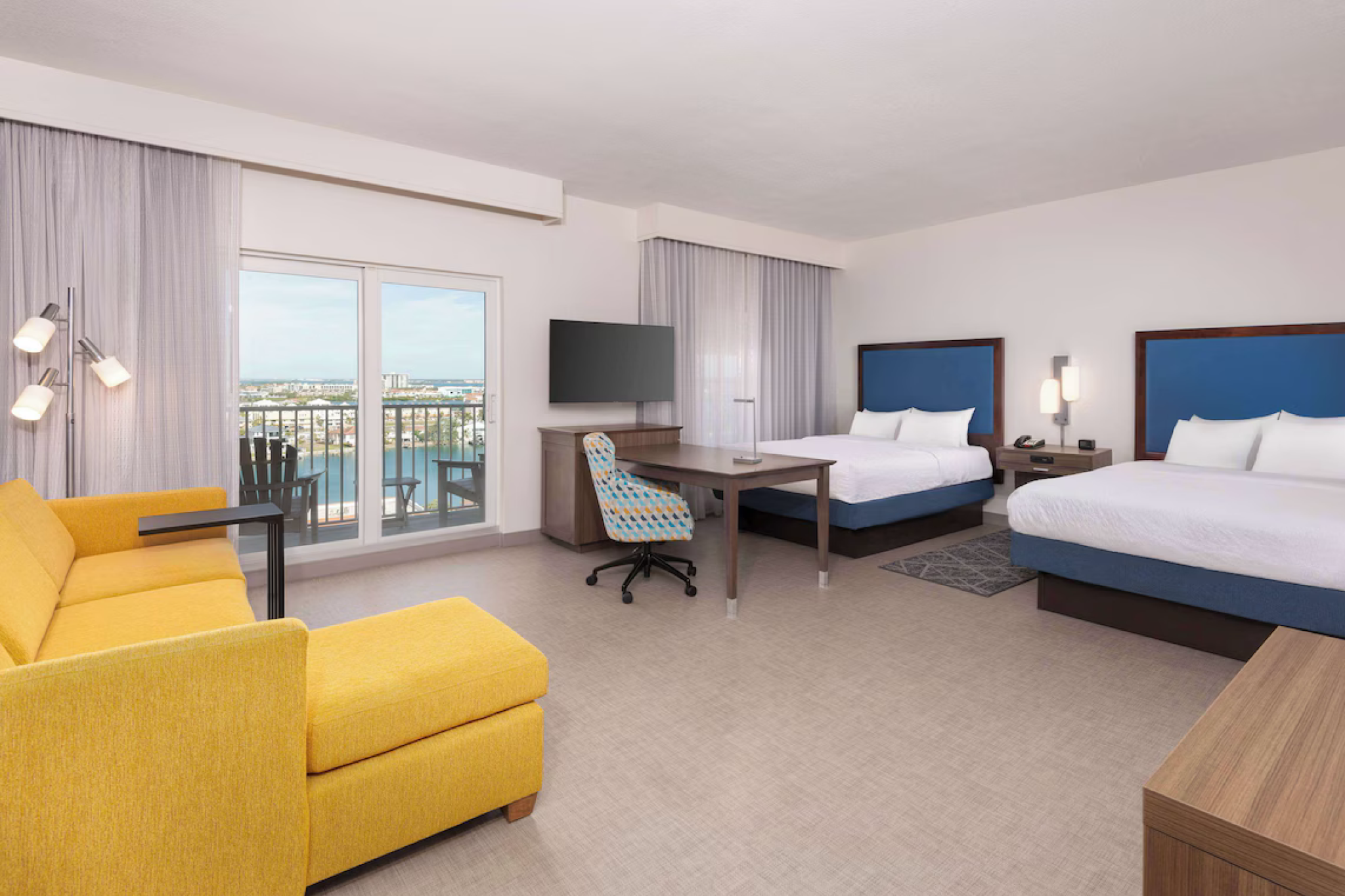 Spacious guest room at Hampton Inn & Suites Clearwater Beach with two queen beds, a yellow sectional sofa, a balcony, and water views through sliding glass doors.