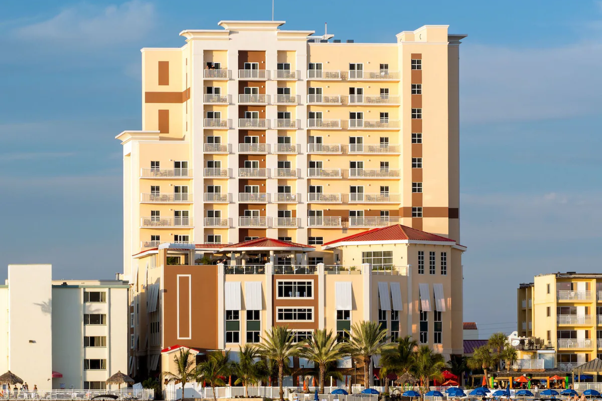 Exterior view of Hampton Inn & Suites Clearwater Beach showing the multi-story building with guest balconies, palm trees, and beachfront access in golden afternoon light.