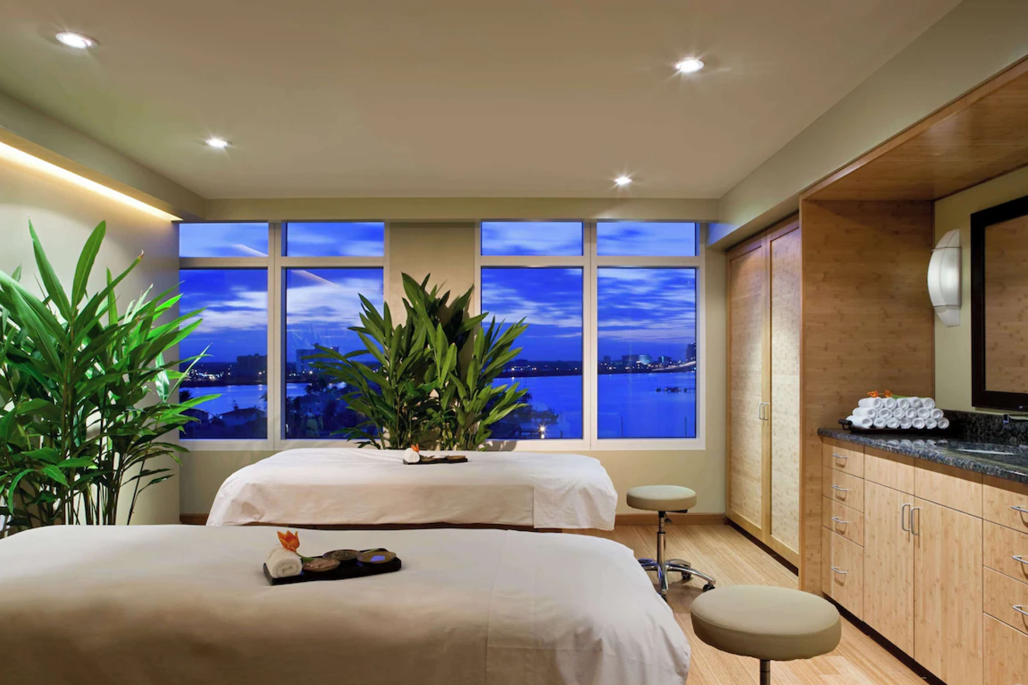 A serene spa treatment room at Hyatt Regency Clearwater Beach Resort and Spa featuring two massage tables, bamboo wood finishes, leafy green plants, and a large window overlooking the calm waters at dusk.