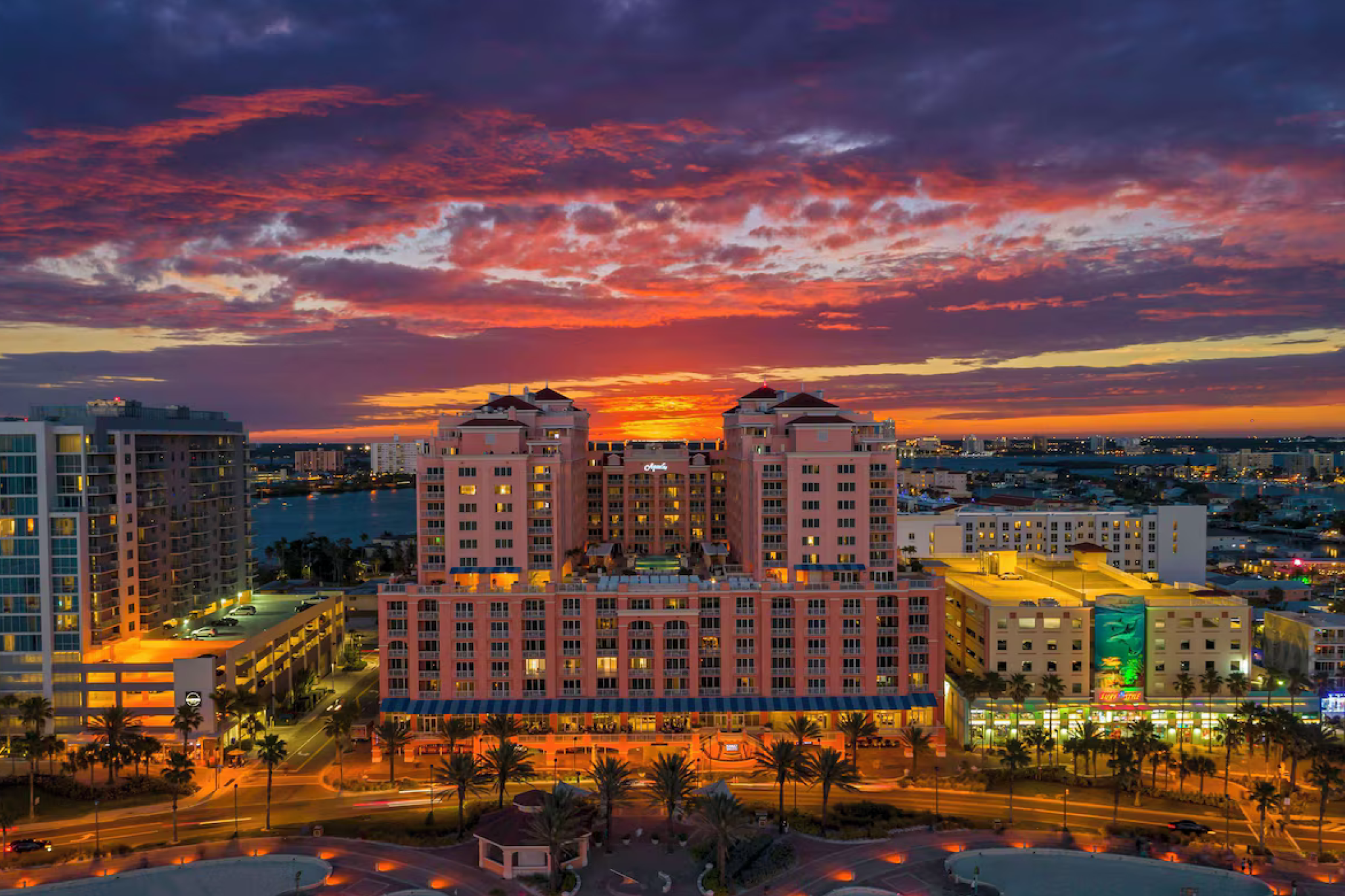 A wide-angle view of the Hyatt Regency Clearwater Beach Resort and Spa at sunset, with the sky painted in dramatic shades of orange, purple, and red, highlighting the pink-hued exterior of the building centered between surrounding city structures.