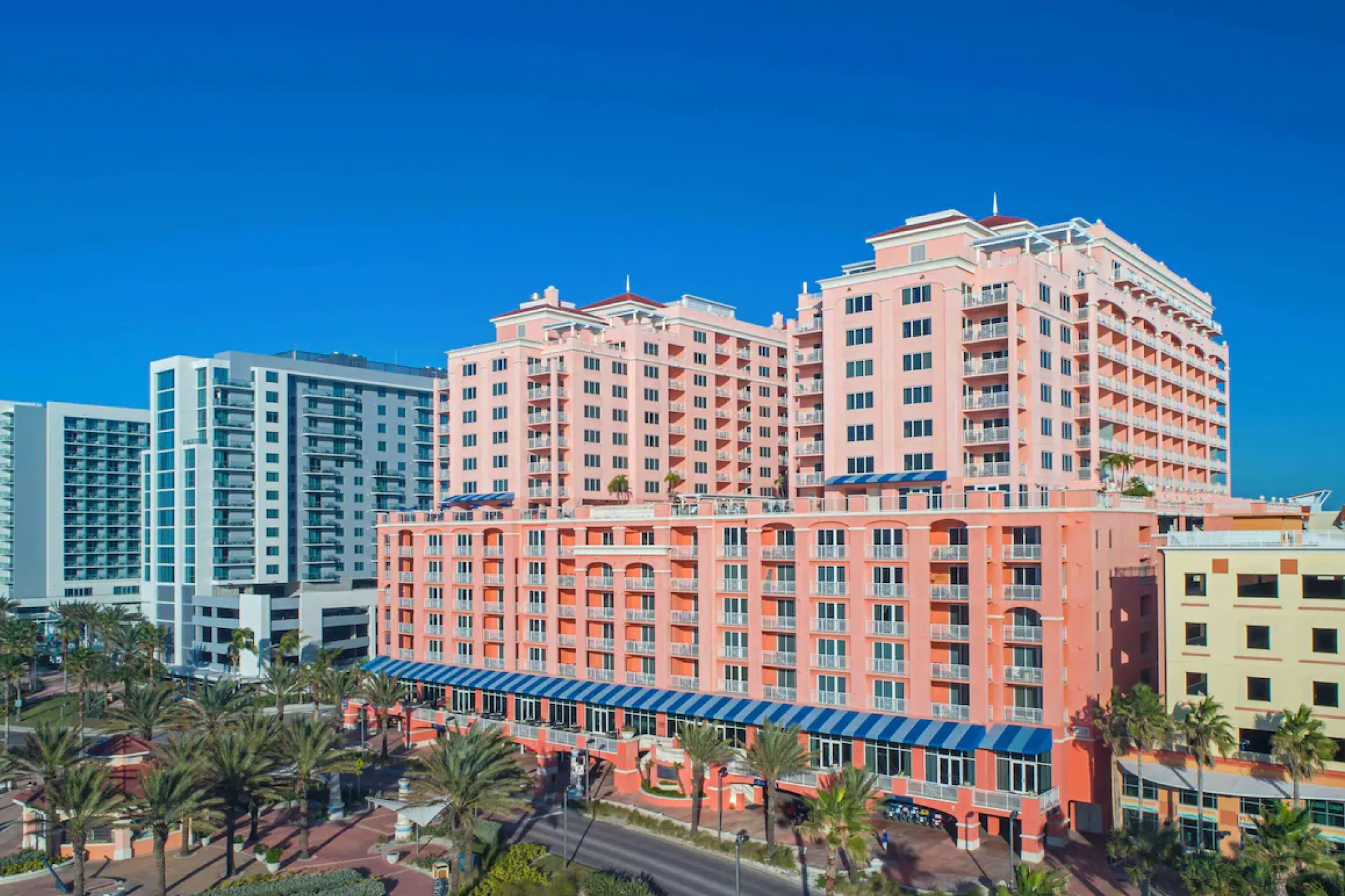 A bright daytime view of the Hyatt Regency Clearwater Beach Resort and Spa, showcasing its multi-story salmon-pink façade with blue awnings, palm-lined streets, and nearby high-rise buildings under a clear blue sky.