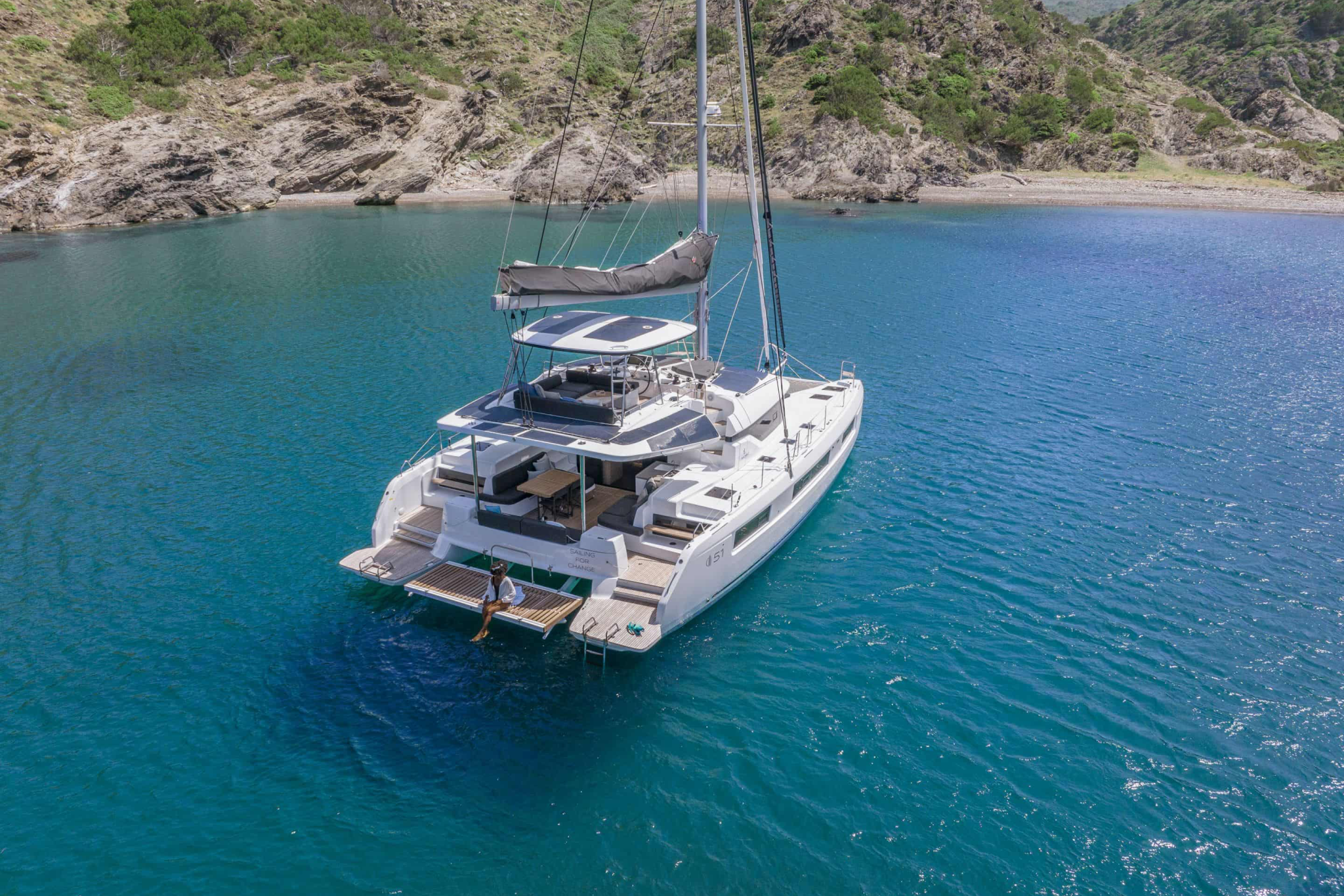 A rear view of the Lagoon 51 anchored in a calm bay, showcasing its wide swim platforms, flybridge, and spacious outdoor living areas.