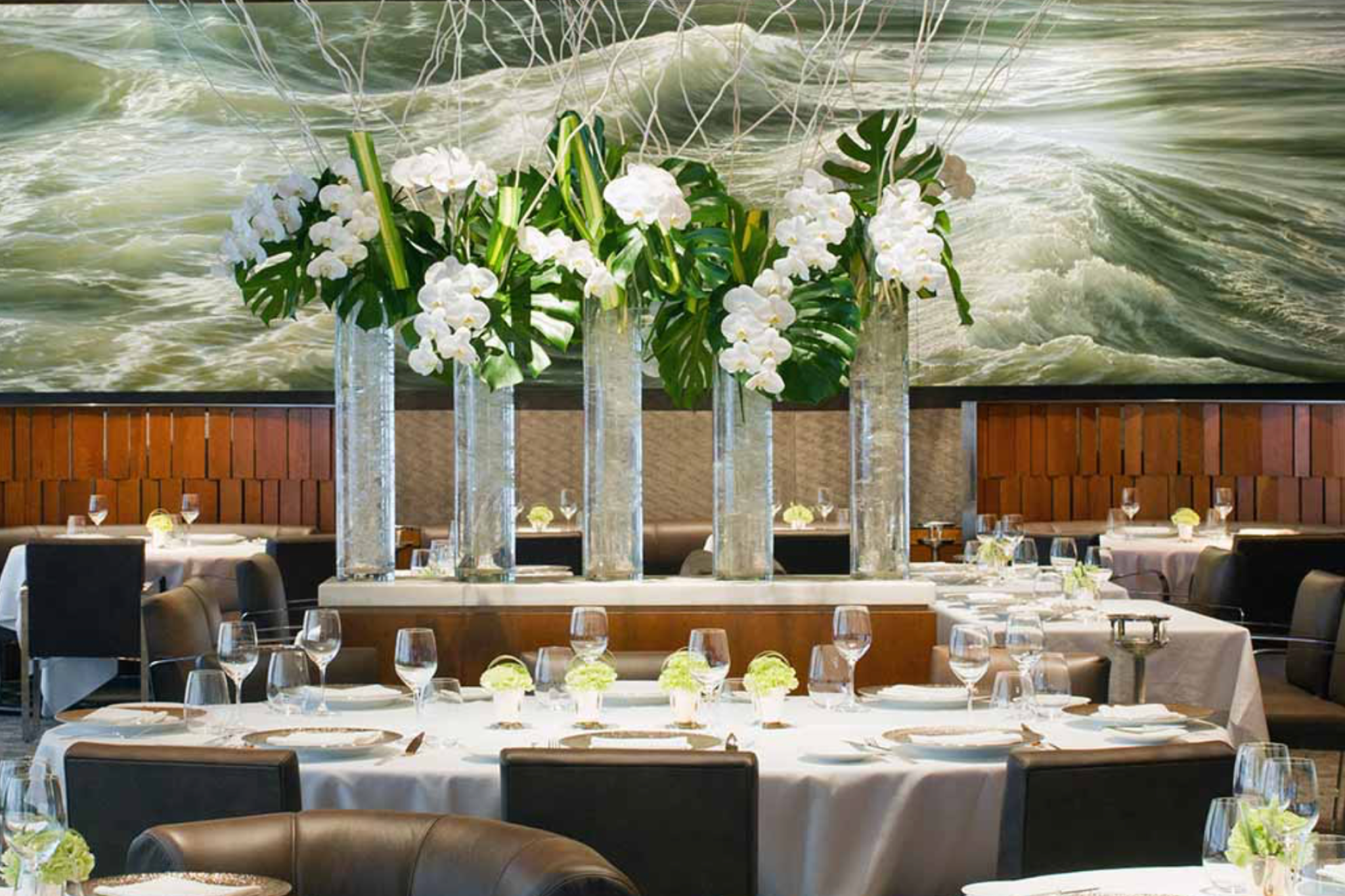 Le Bernardin’s main dining area featuring dramatic floral arrangements and large-scale ocean artwork behind round tables.