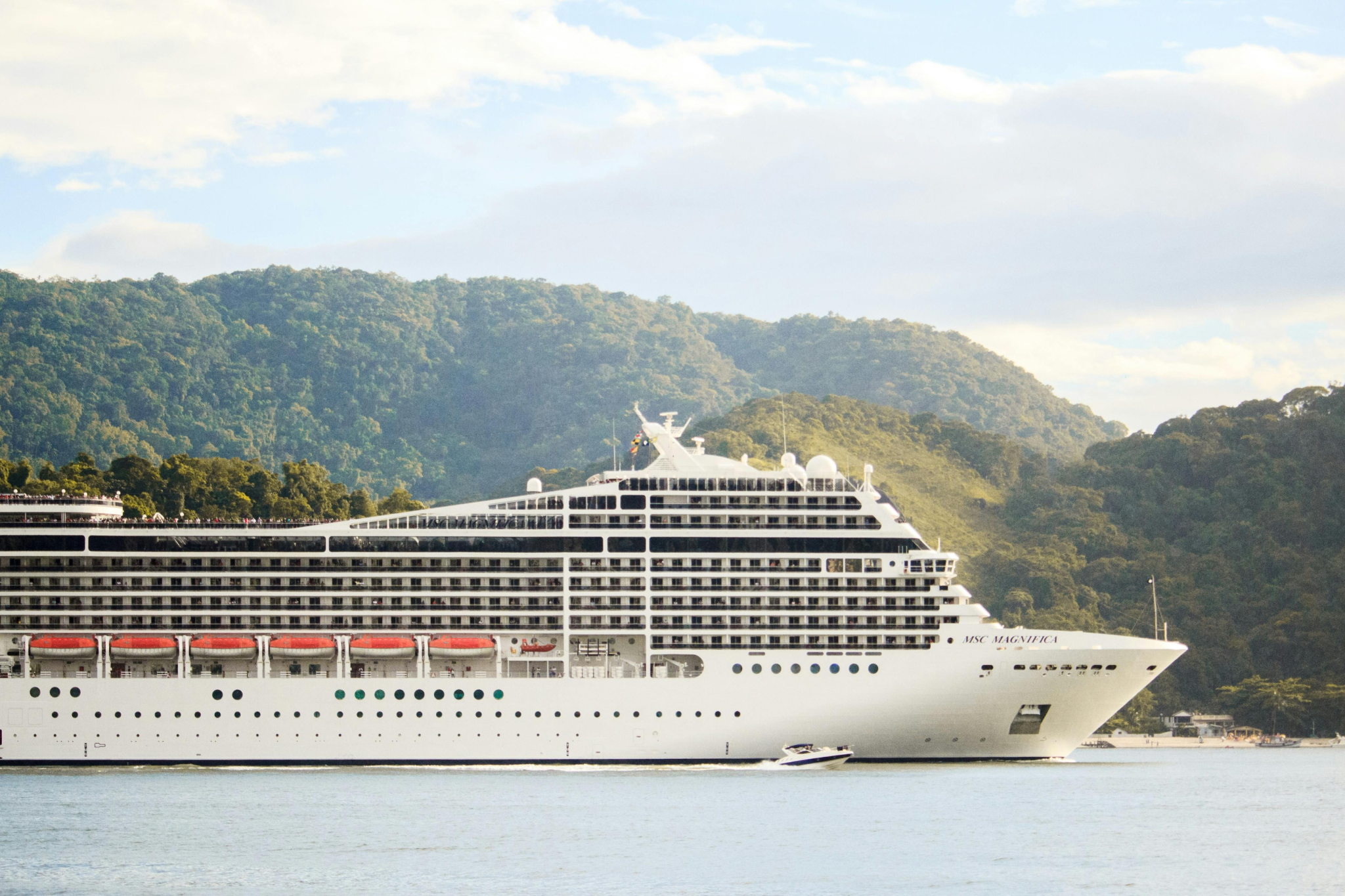 MSC Magnifica cruise ship sailing along a lush, green coastline with a scenic mountain backdrop.