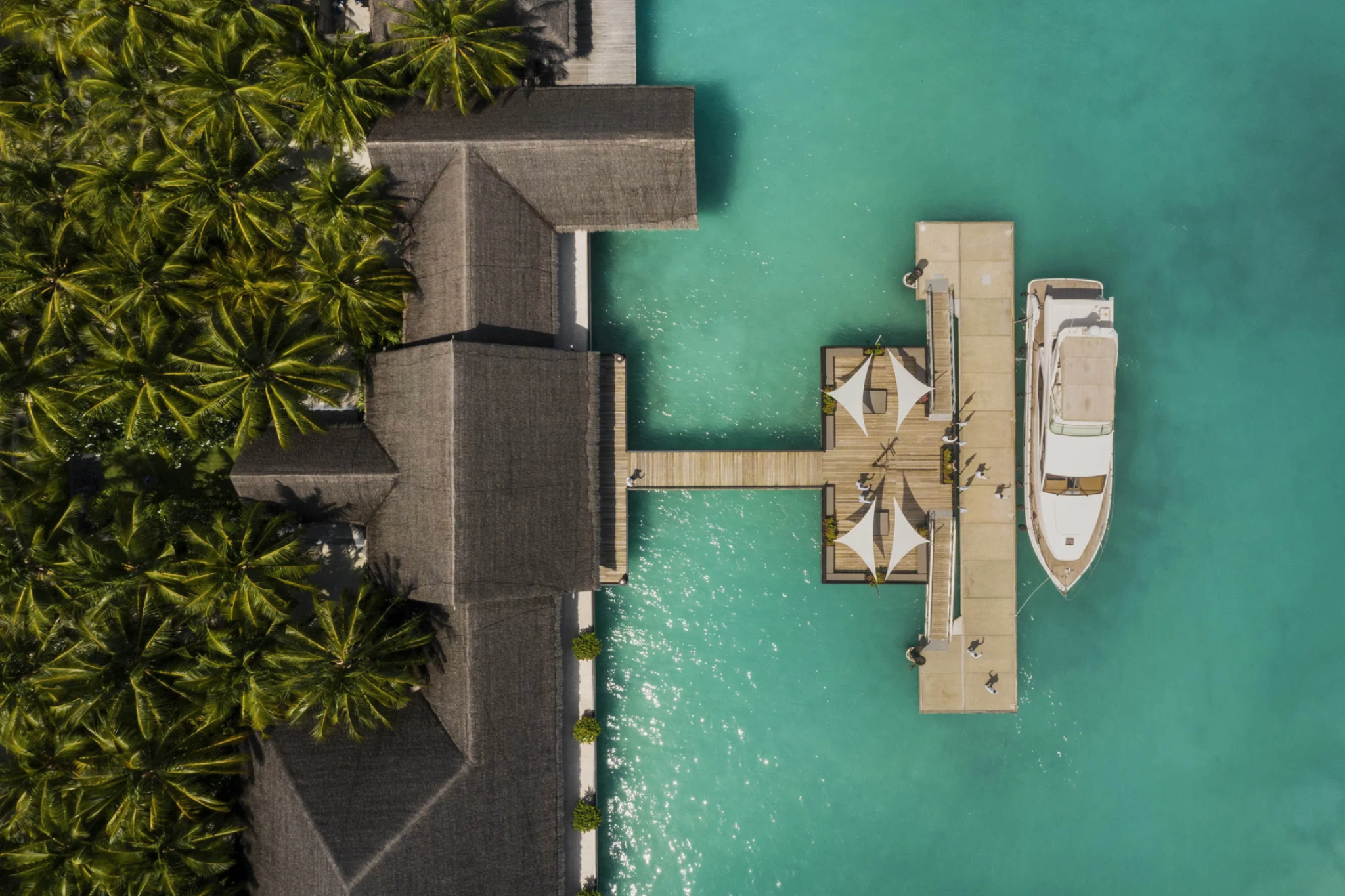Yacht docked at the arrival pier of One&Only Reethi Rah beside a thatched-roof welcome lounge and palm-covered shoreline.