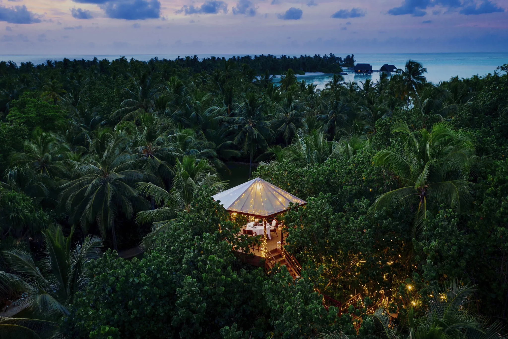 Private jungle dining pavilion at One&Only Reethi Rah lit with string lights at sunset, surrounded by lush tropical greenery.