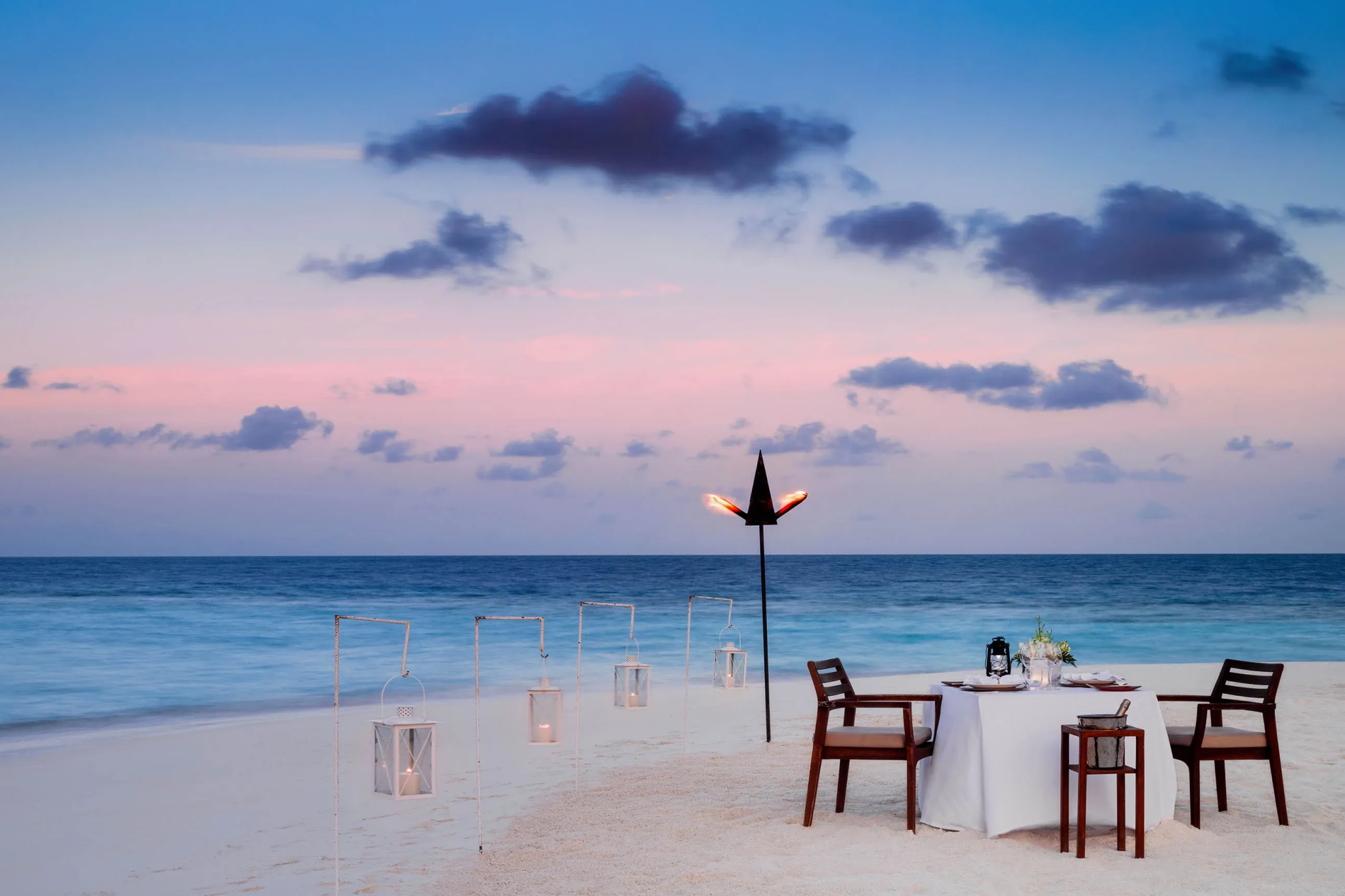 Romantic dining setup on the beach at One&Only Reethi Rah, with a lantern-lit table facing the sea during sunset.