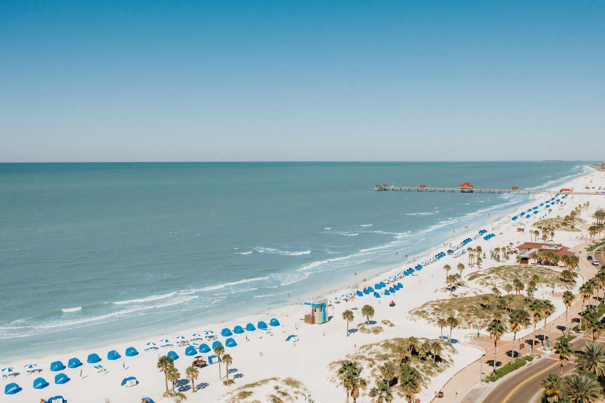 A stunning aerial view of Clearwater Beach with rows of blue beach umbrellas and a clear sightline to Pier 60 near Opal Sands Resort.