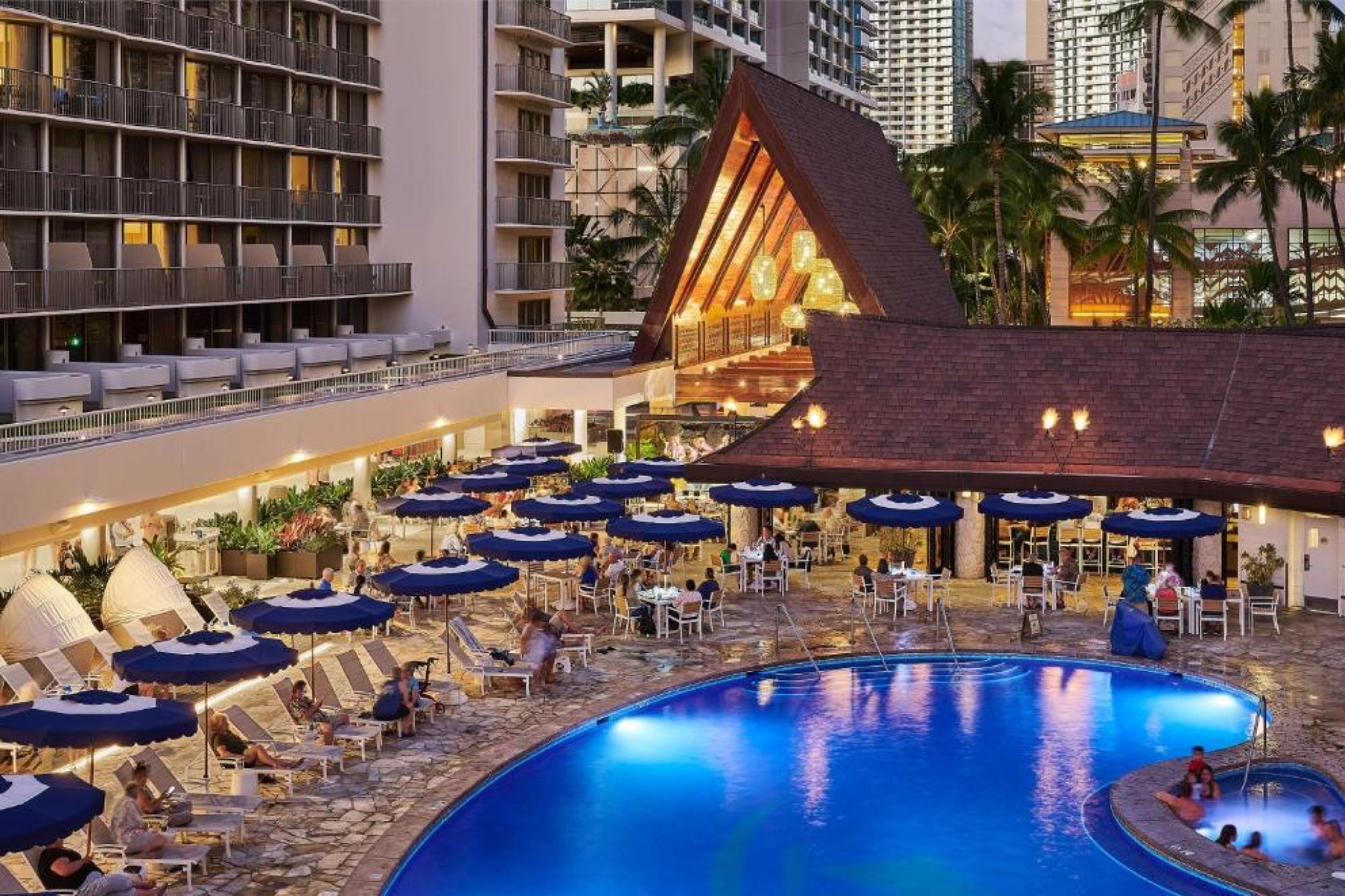 Evening poolside scene at Outrigger Reef Waikiki Beach Resort with guests dining at Kani Ka Pila Grille under blue umbrellas beside the illuminated pool.