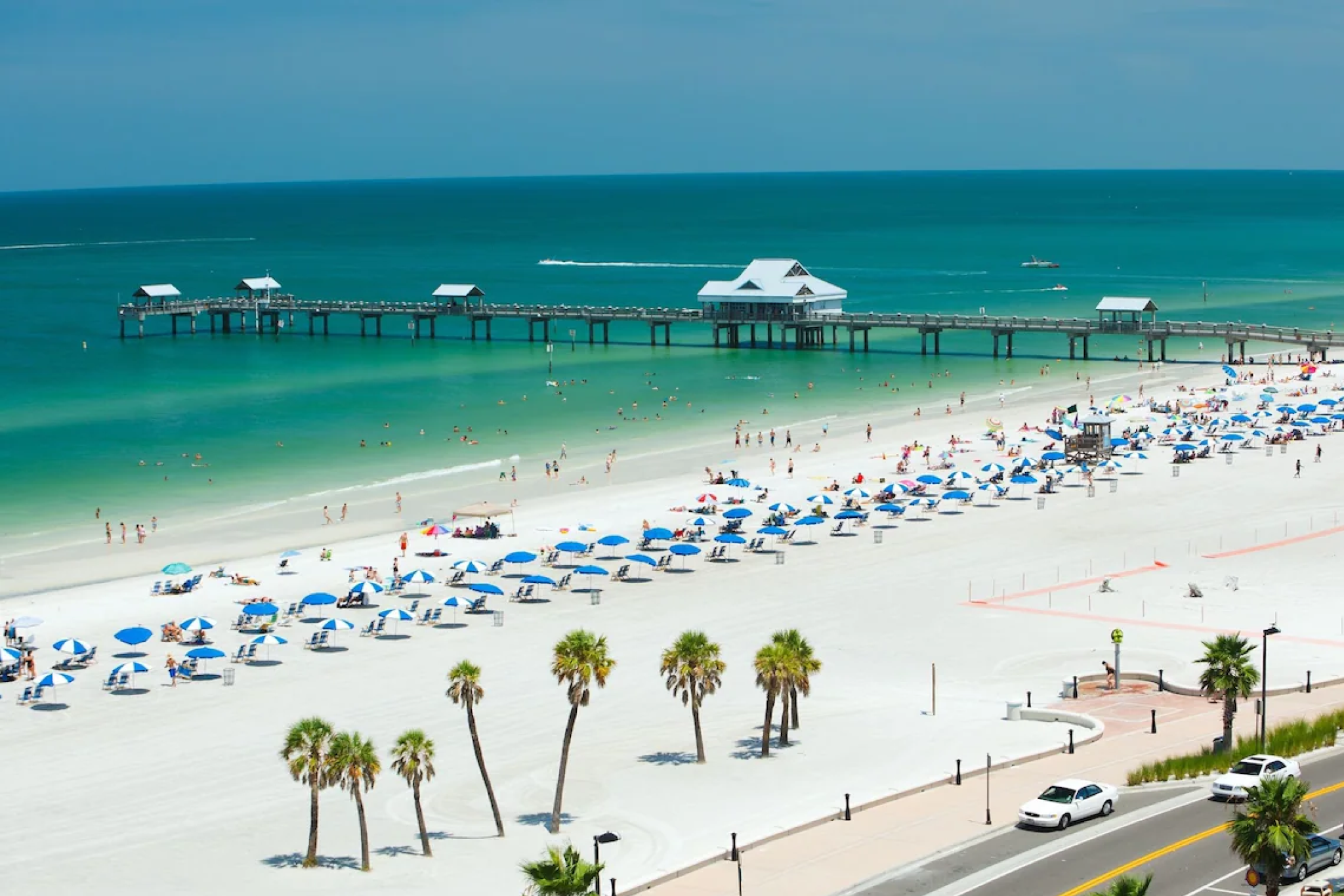 Wide aerial view of Pier 60 and Clearwater Beach, showing the turquoise Gulf waters, beach umbrellas, and guests enjoying the sun near Pier House 60 Clearwater Beach Marina Hotel.