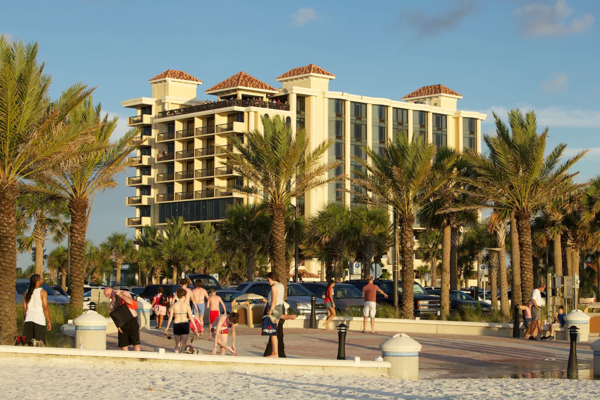 Street-level view of Pier House 60 Clearwater Beach Marina Hotel with palm trees and beachgoers walking along the nearby sandy path.