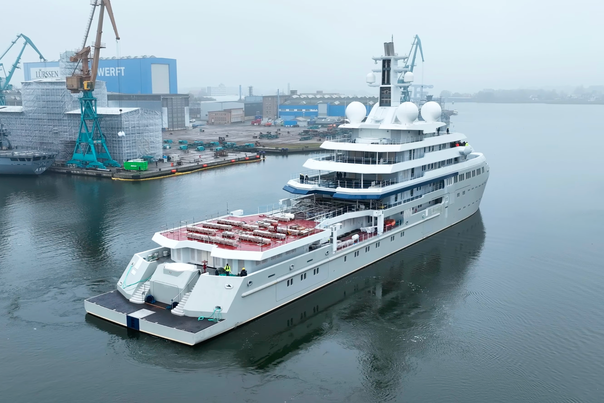 Project Shackleton superyacht seen from the stern during yard maneuvers, with scaffolding and shipyard cranes in the background, showing outfitting progress ahead of sea trials and delivery.