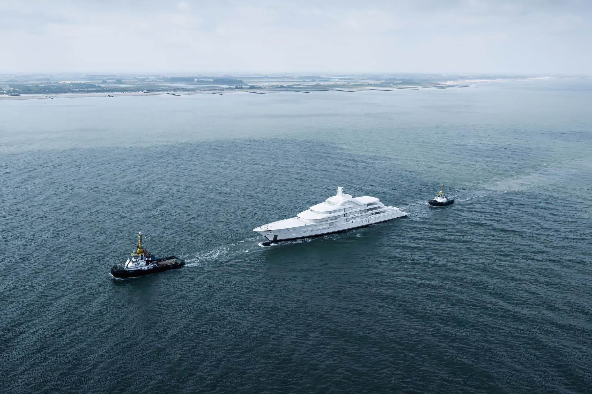 Aerial view of Project Tanzanite under tow between support vessels, highlighting its immense scale, streamlined hull form, and displacement design as the yacht transitions toward sea trials.