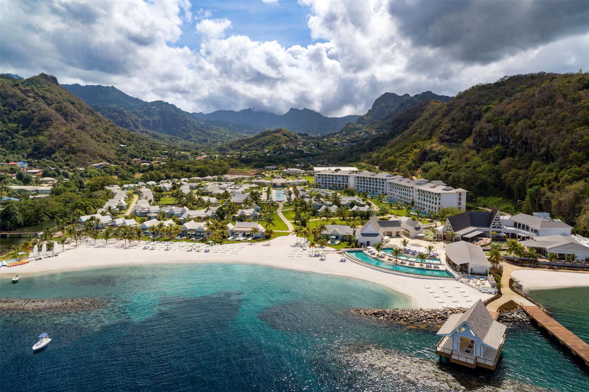 Aerial view of Sandals Saint Vincent showing the white-sand beach, turquoise bay, beachfront villas, infinity pool, and the surrounding rainforest-covered mountains.