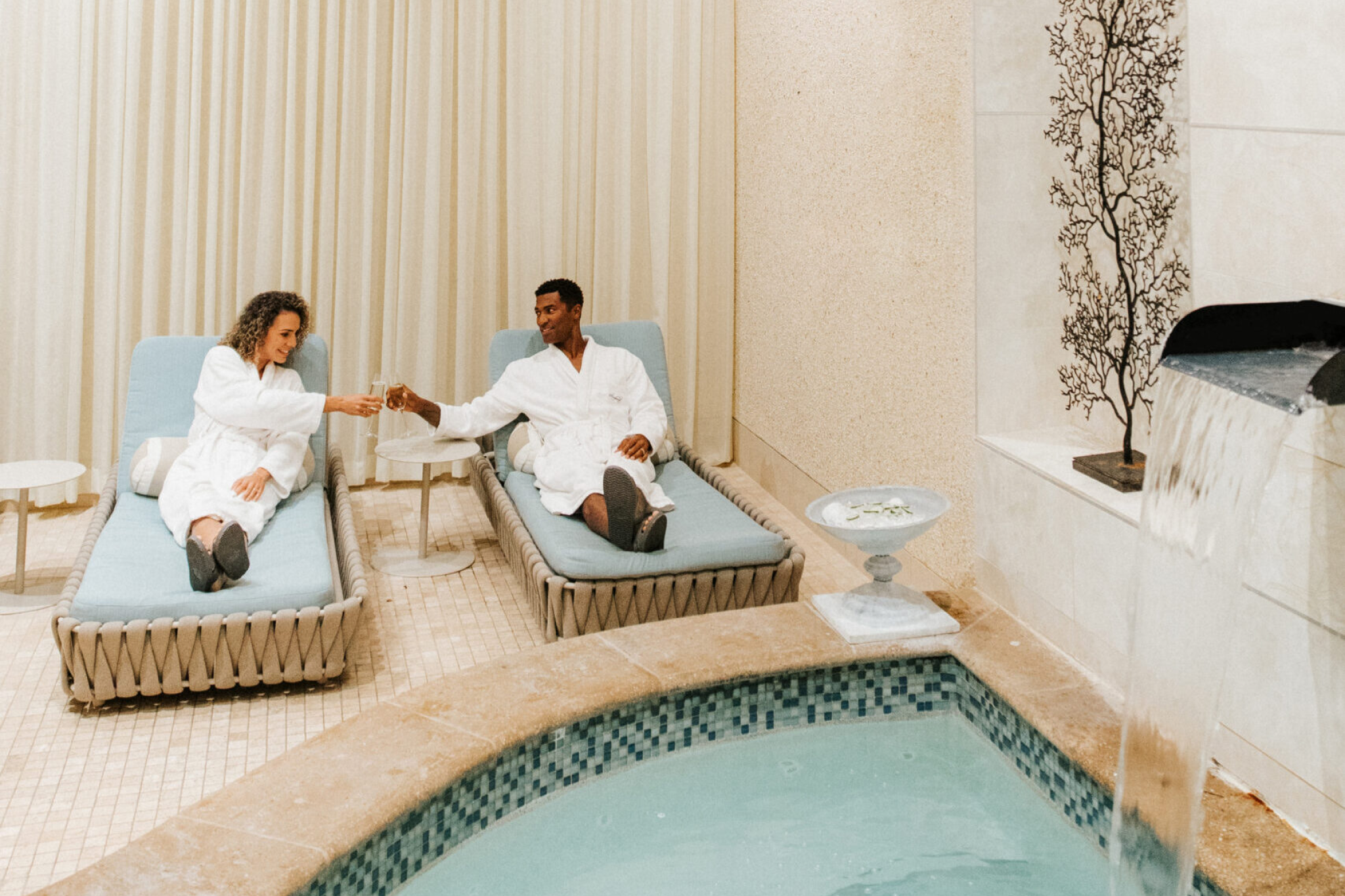 Couple relaxing on loungers and toasting champagne beside a tranquil indoor water feature at the luxurious Opal Spa inside Sandpearl Resort.