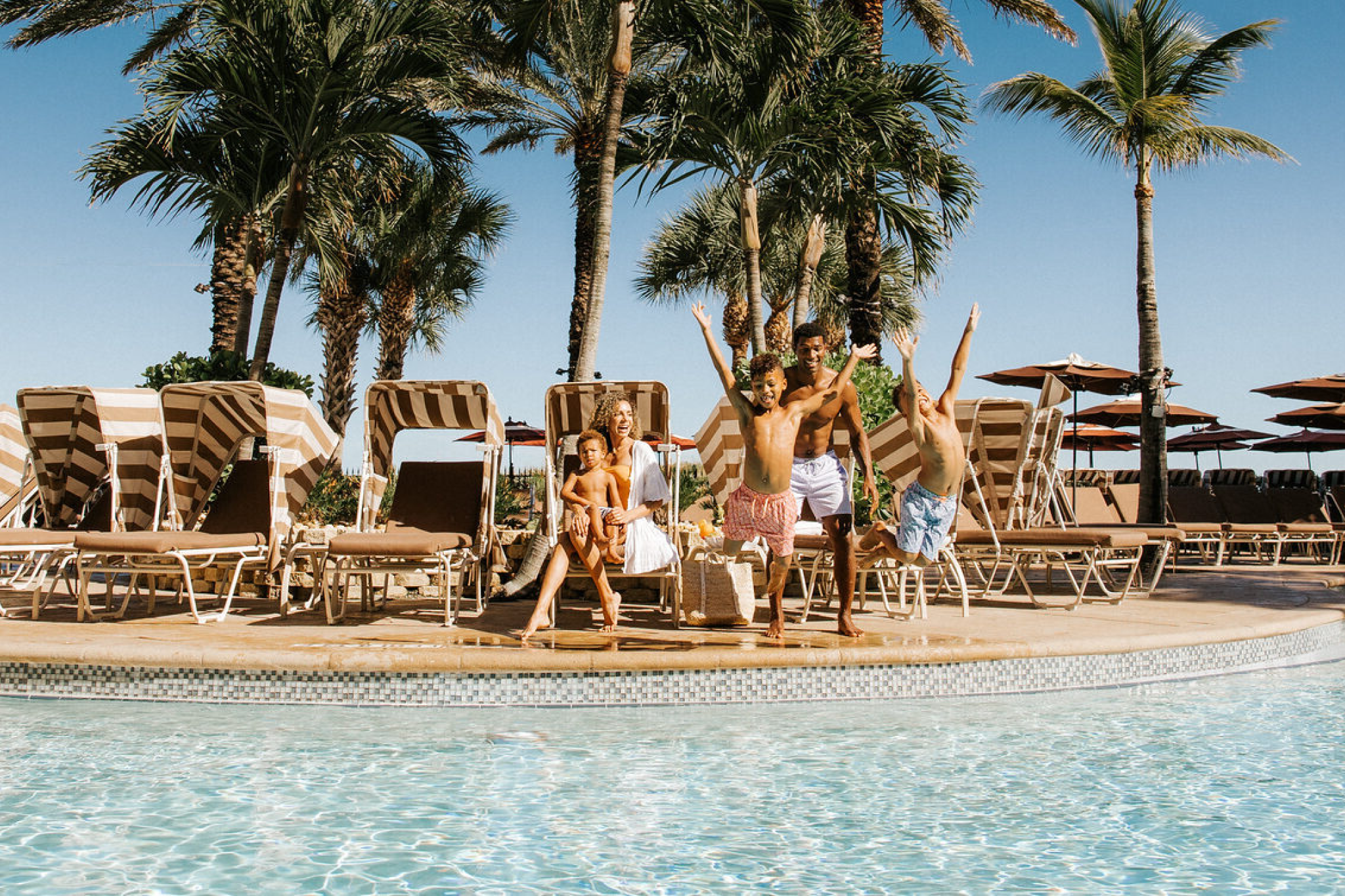 Family enjoying a sunny day by the lagoon-style pool at Sandpearl Resort, with kids jumping into the water surrounded by palm trees and cabanas.