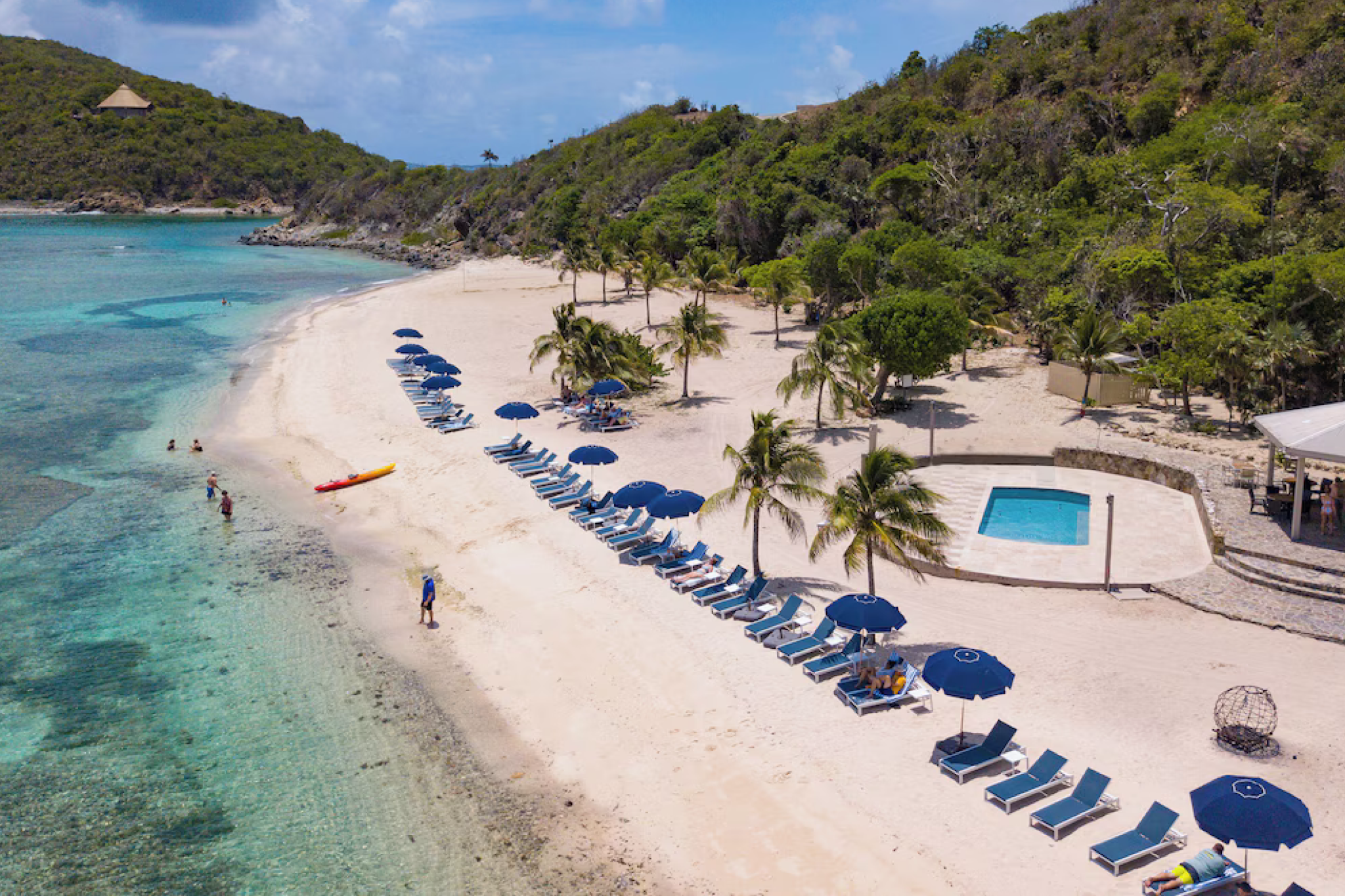 Wide sandy beach at Scrub Island Resort Spa & Marina with rows of blue umbrellas and lounge chairs, calm waters, and nearby hills.