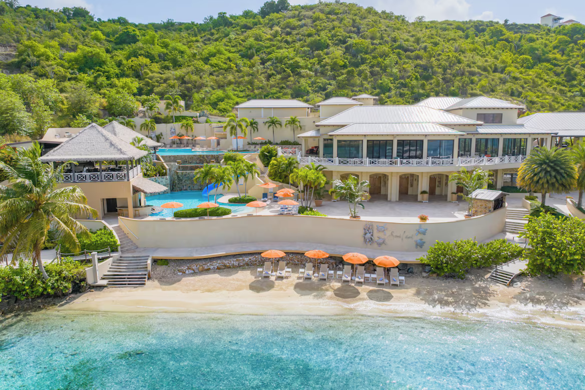 Aerial view of Scrub Island Resort Spa & Marina showing the beachfront, hillside buildings, and cascading infinity pools surrounded by lush green hills.