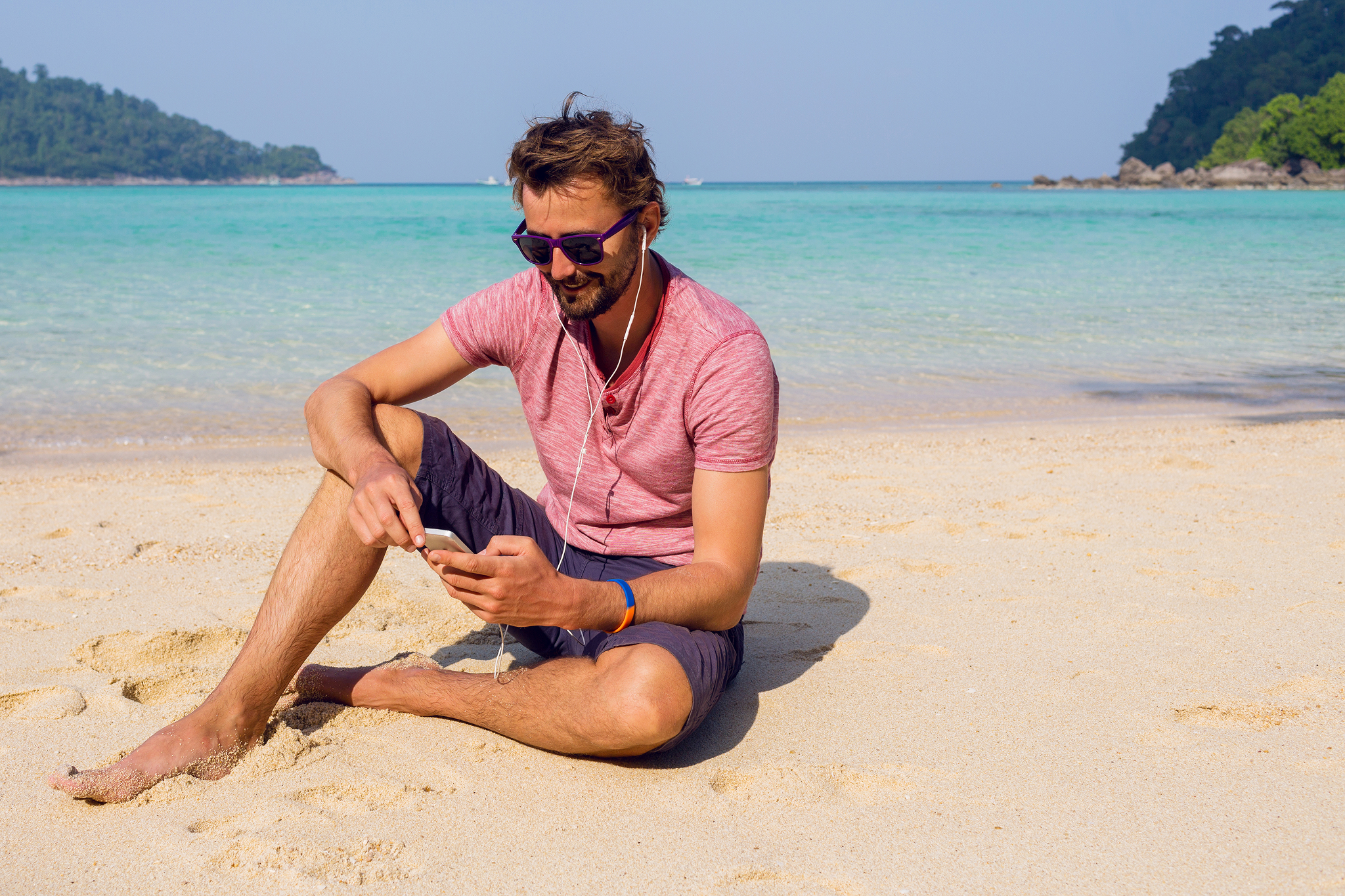 Man sitting on the beach at Scrub Island Resort Spa & Marina, wearing sunglasses and listening to music while looking at his phone.