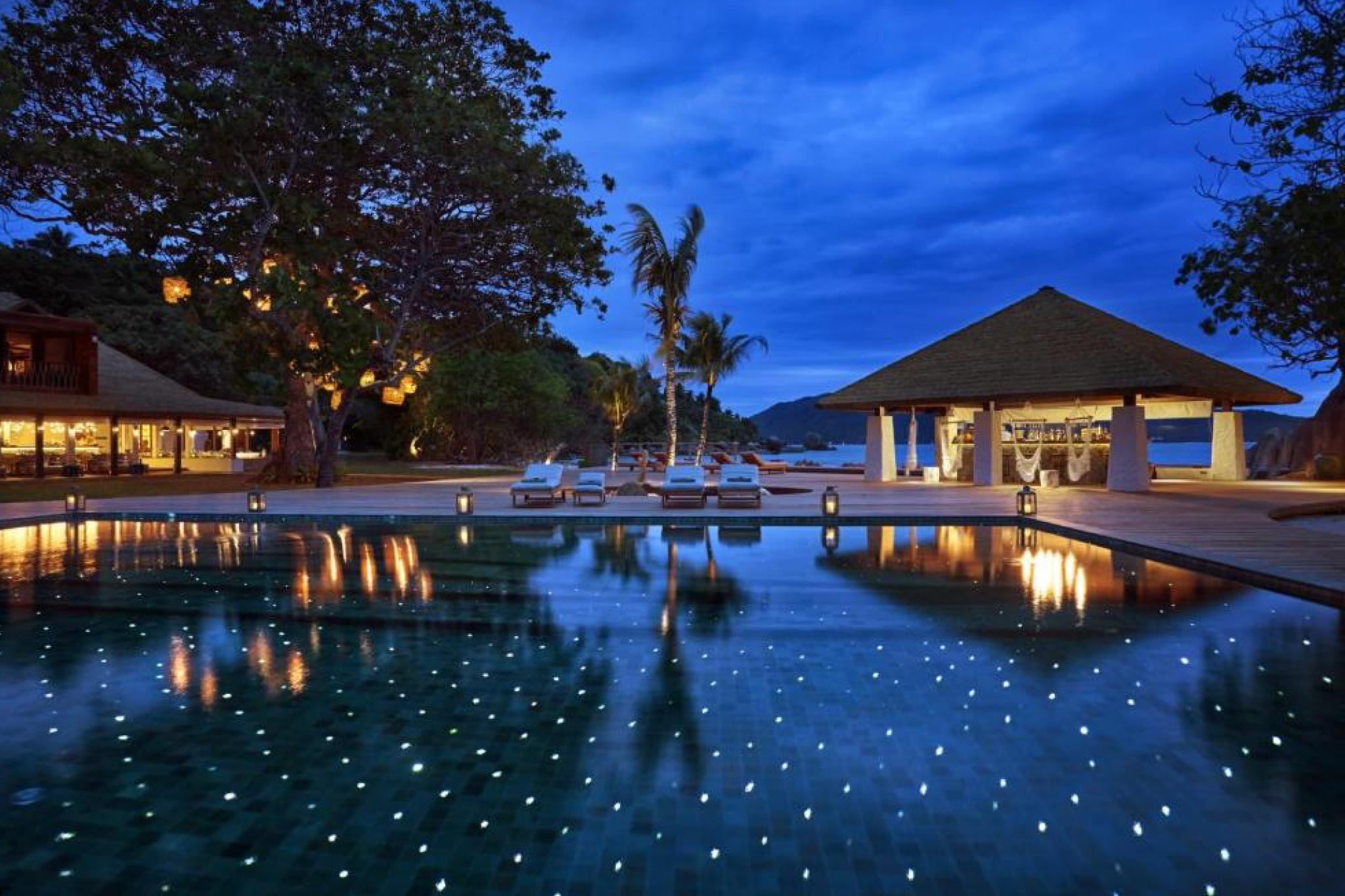 Resort pool at dusk reflecting lantern-lit trees and the poolside bar, with loungers and palm trees under a twilight sky at Six Senses Zil Pasyon.